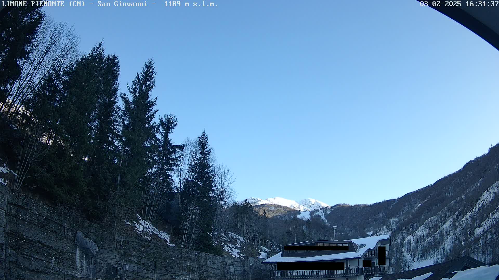 A snow-dusted mountain range is visible under a clear blue sky, behind a building partially obscured by evergreens and a rocky hillside.