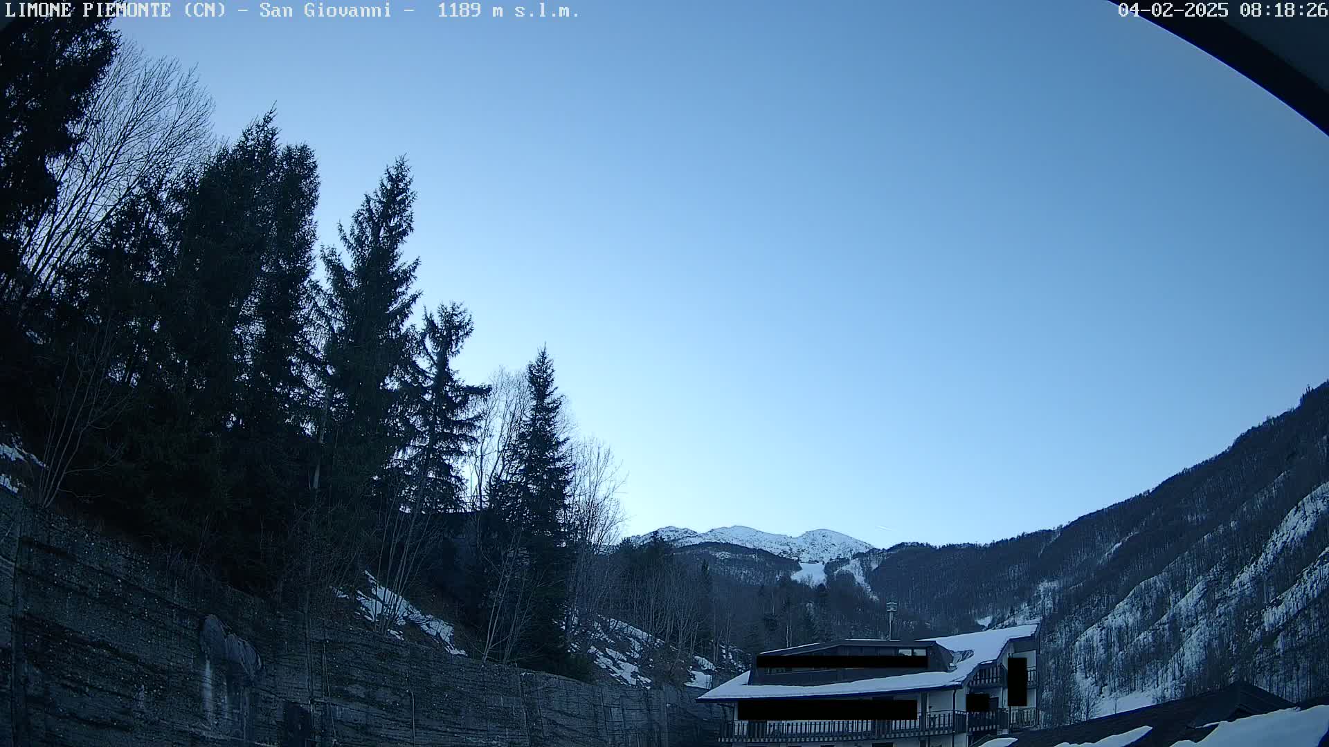 A clear blue sky is over a snowy mountainside with evergreen trees and a building partially visible.