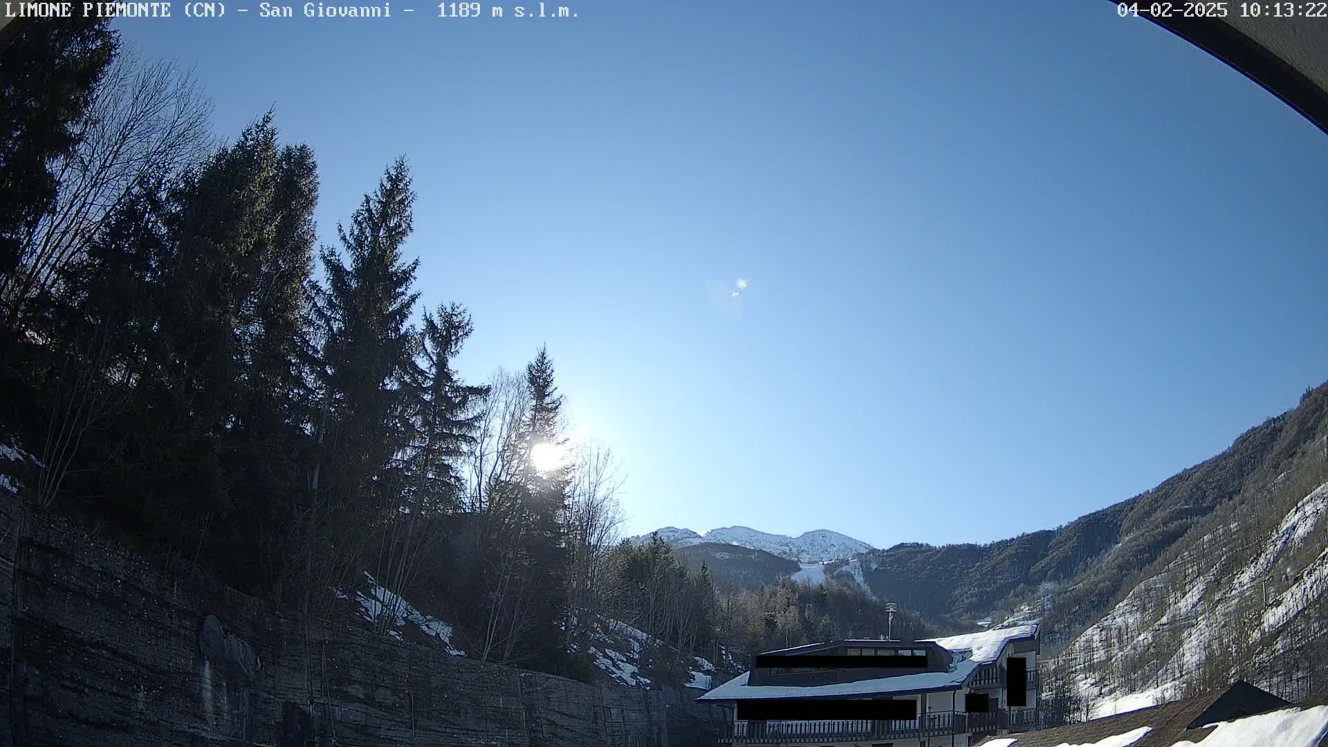 A sunny day in a mountainous area shows a building partially obscured by evergreen trees and snow-covered slopes.