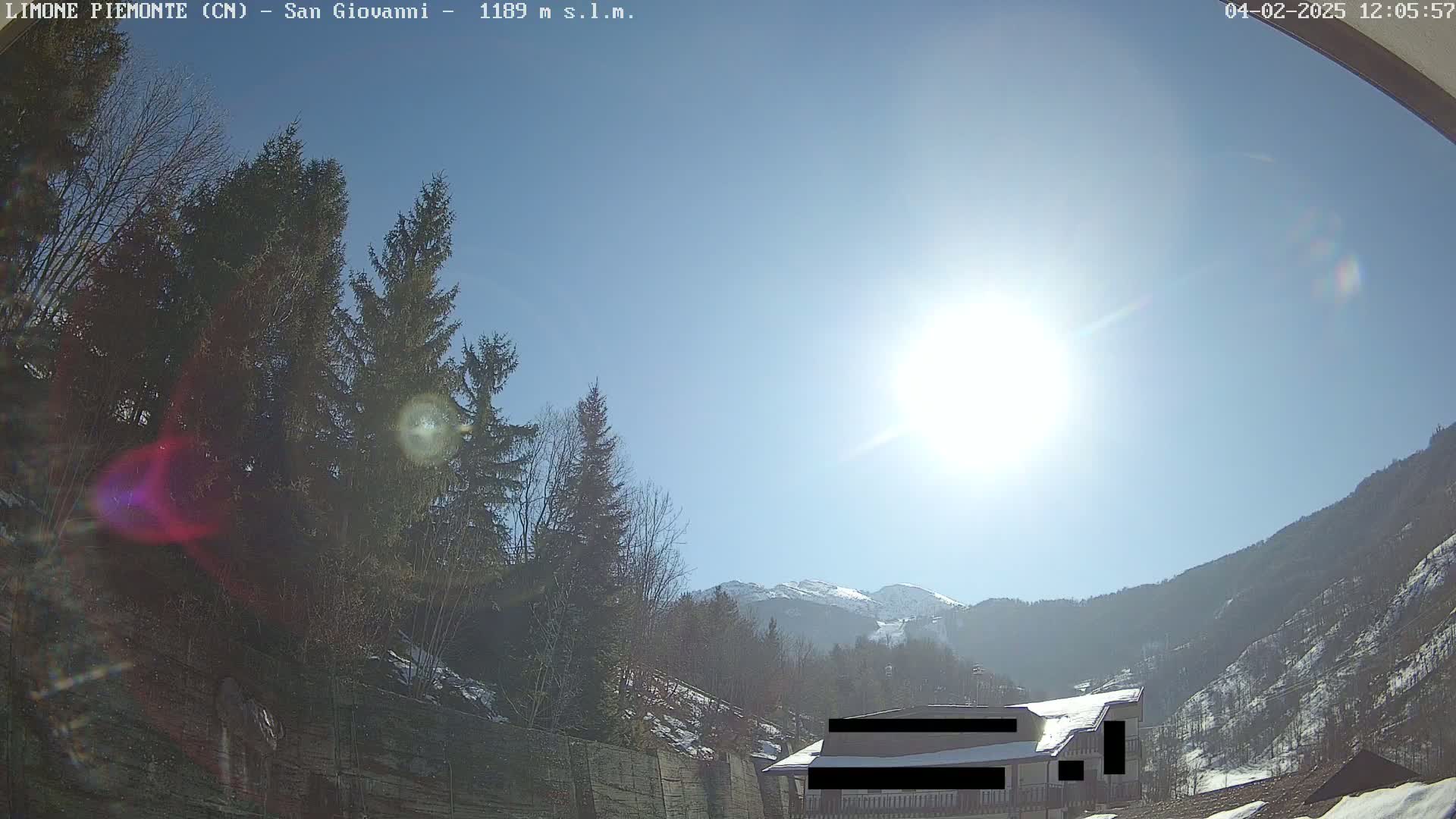 A sunny day reveals a mountainous landscape with evergreen trees, a building partially obscured, and a bright sun in a clear blue sky.