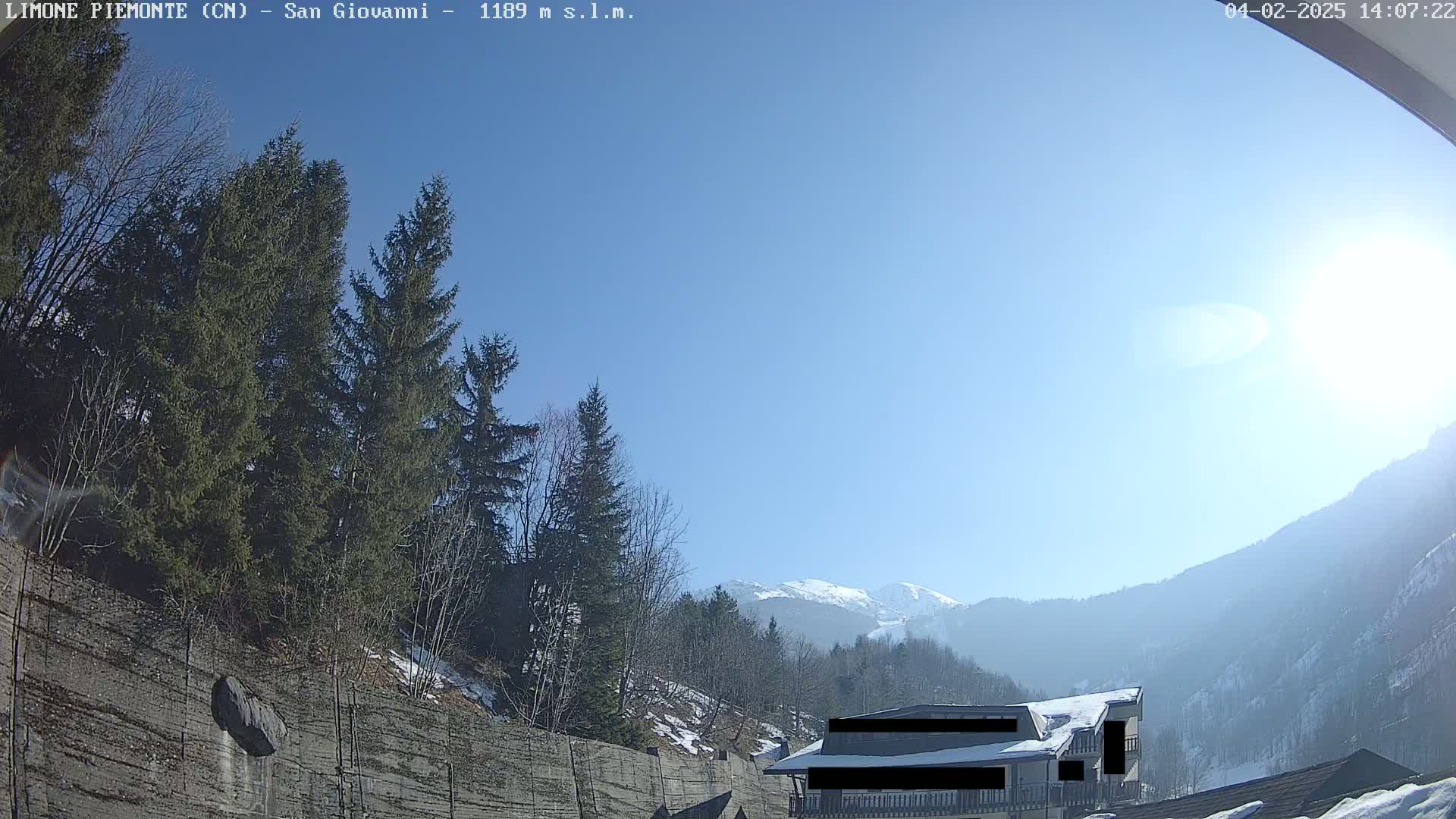 A sunny day in a mountainous region shows evergreen trees, a building partially obscured, and snow-capped mountains in the distance.