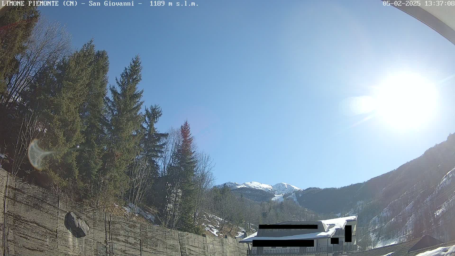 A sunny day reveals a snow-capped mountain in the distance, behind a building partially obscured by evergreens and a retaining wall.