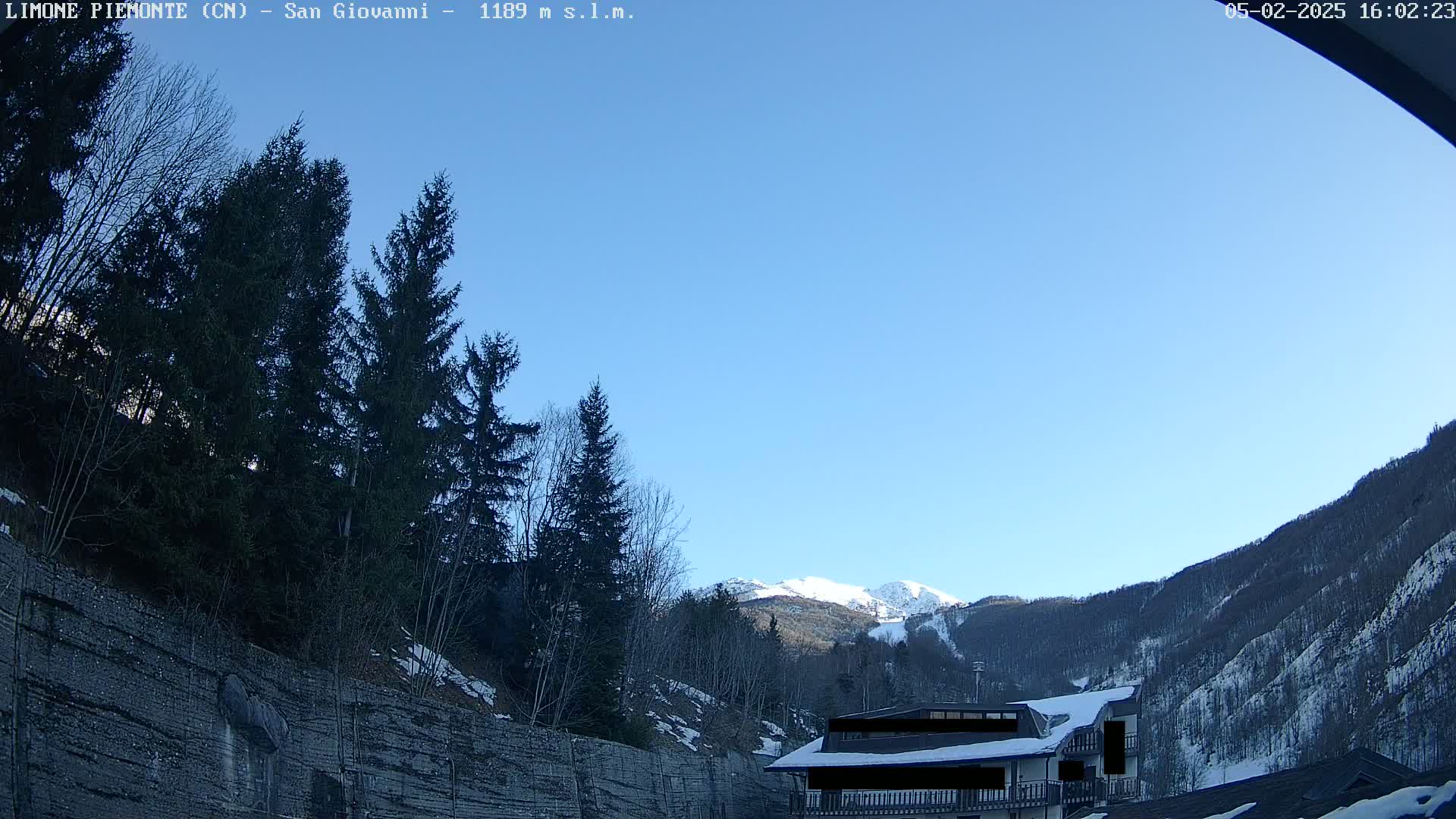 A clear blue sky overlooks snow-dusted mountains and evergreen trees, with a building partially visible in the foreground.