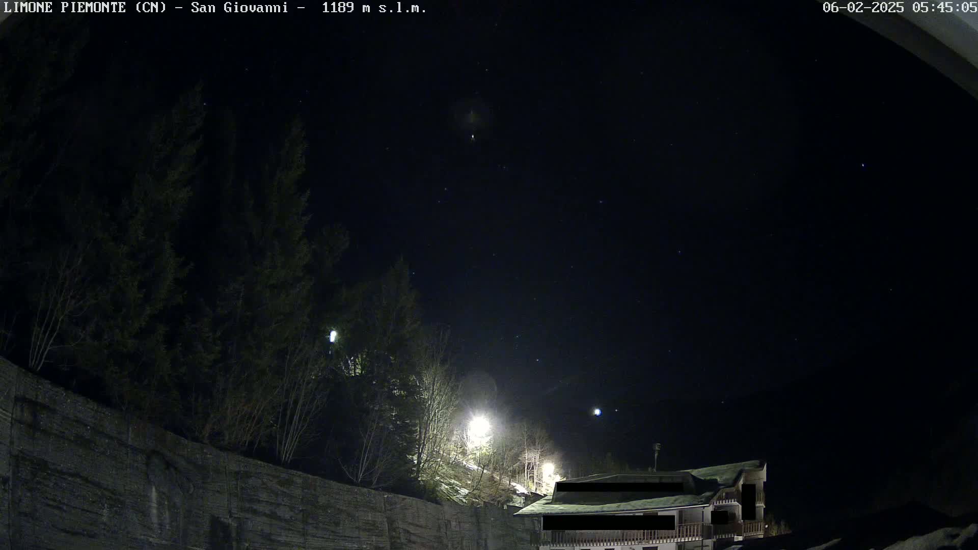 A night scene shows a dark, starlit sky over a snow-dusted hillside with dark evergreen trees and a house illuminated by streetlights.