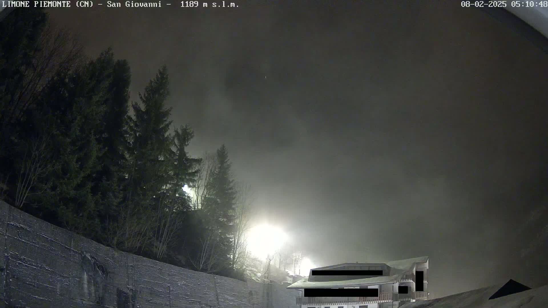 A nighttime scene of fog obscuring a building and evergreen trees behind a snow-covered wall, illuminated by artificial lights.