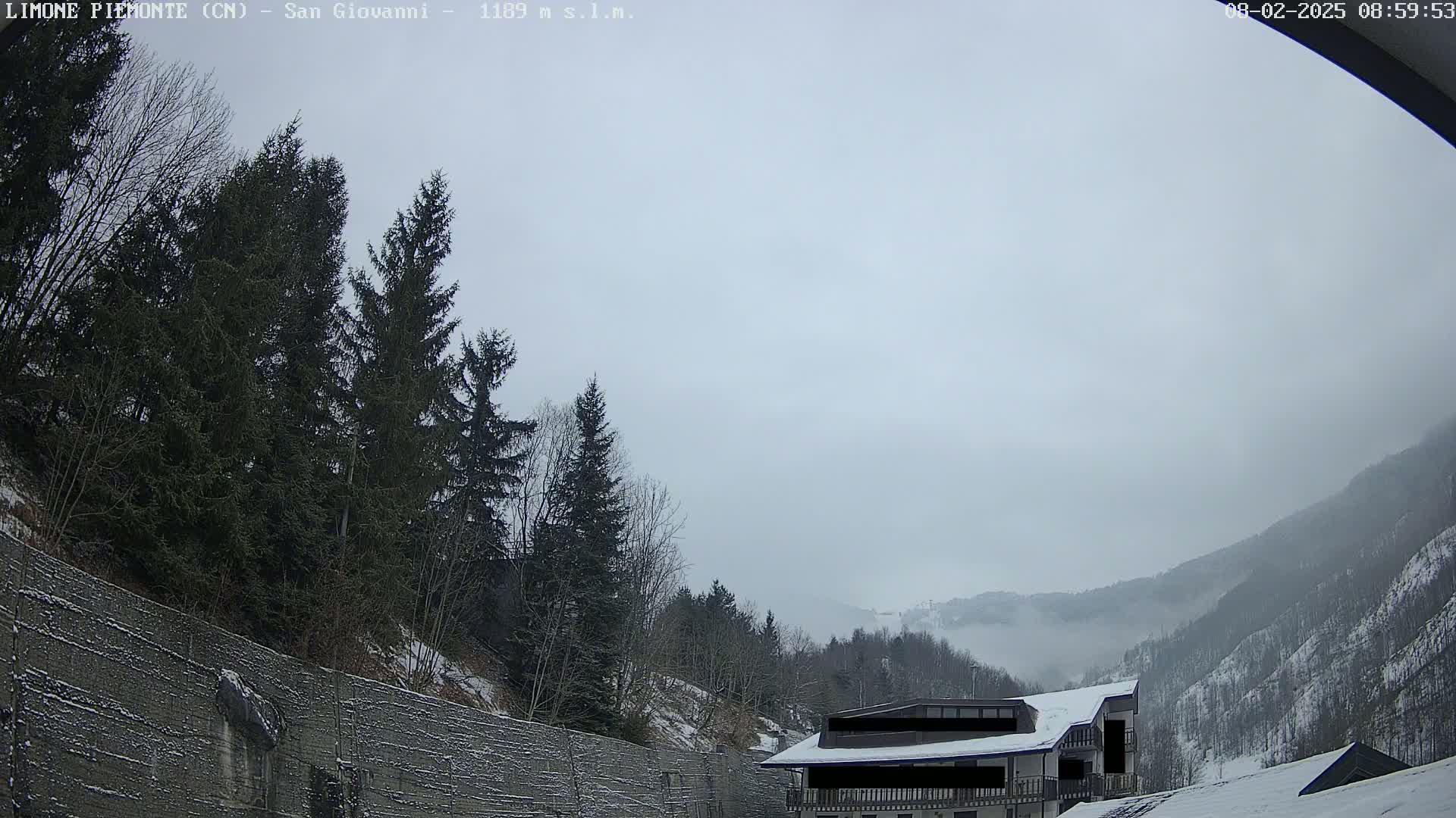 A snow-covered mountain valley scene on an overcast day shows a building near a retaining wall, with evergreens on a hillside in the foreground.