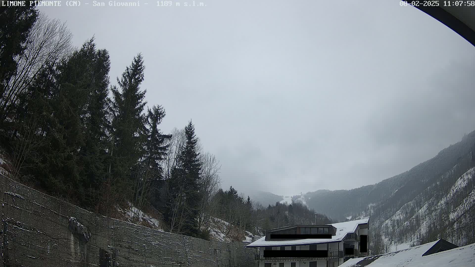 A cloudy, snowy mountain landscape is seen from behind a tall retaining wall, with evergreens in the foreground and a building partially visible in the mid-ground.