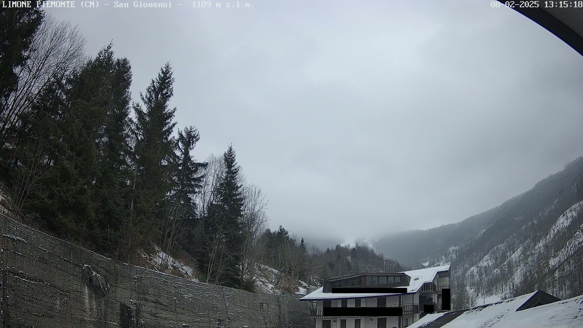 A snow-covered mountain valley scene under an overcast sky shows evergreen trees, a building, and a retaining wall.