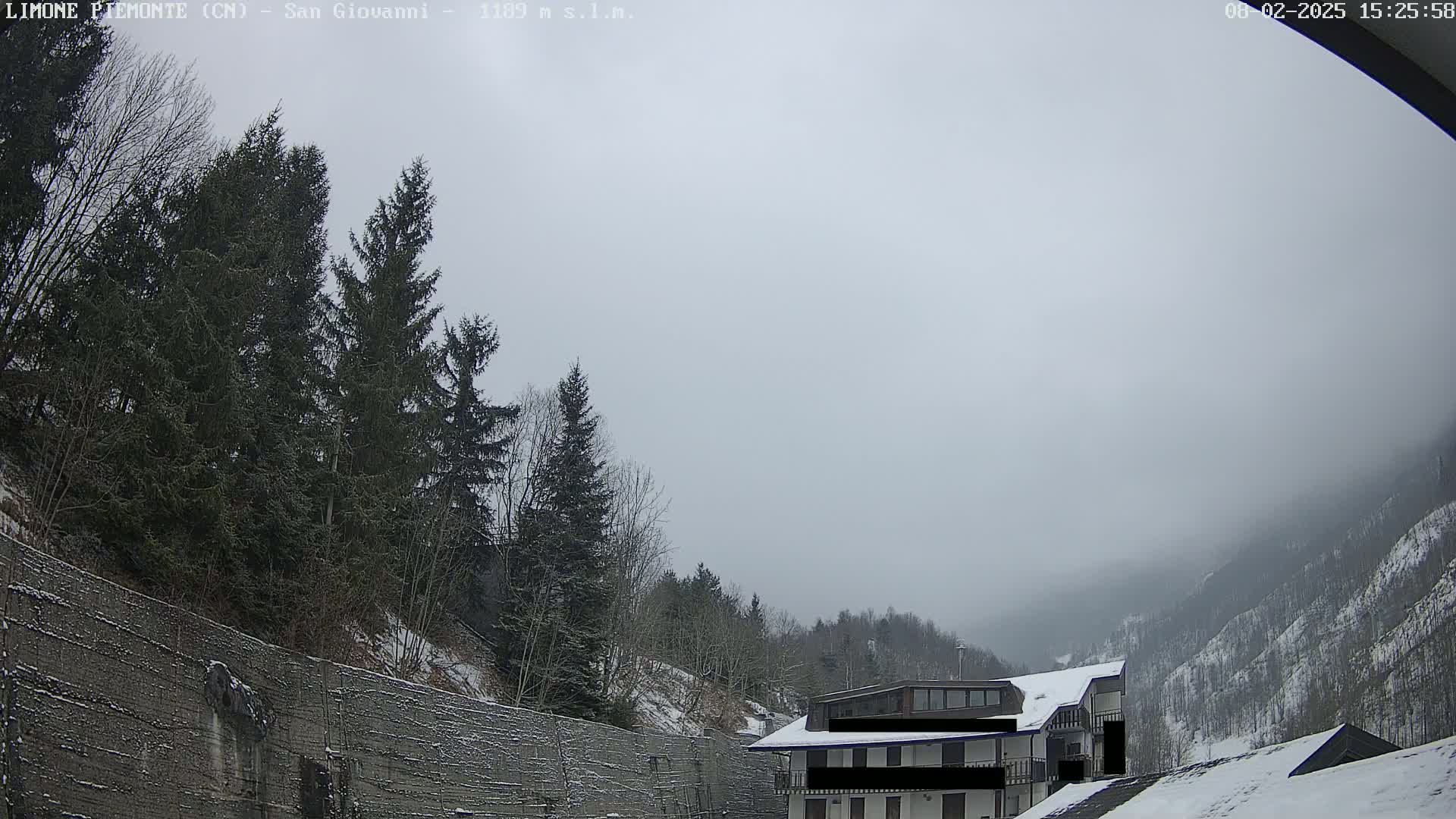 A snow-covered mountain valley is visible under an overcast sky, with evergreen trees lining a concrete wall in the foreground and a multi-story building partially visible in the midground.