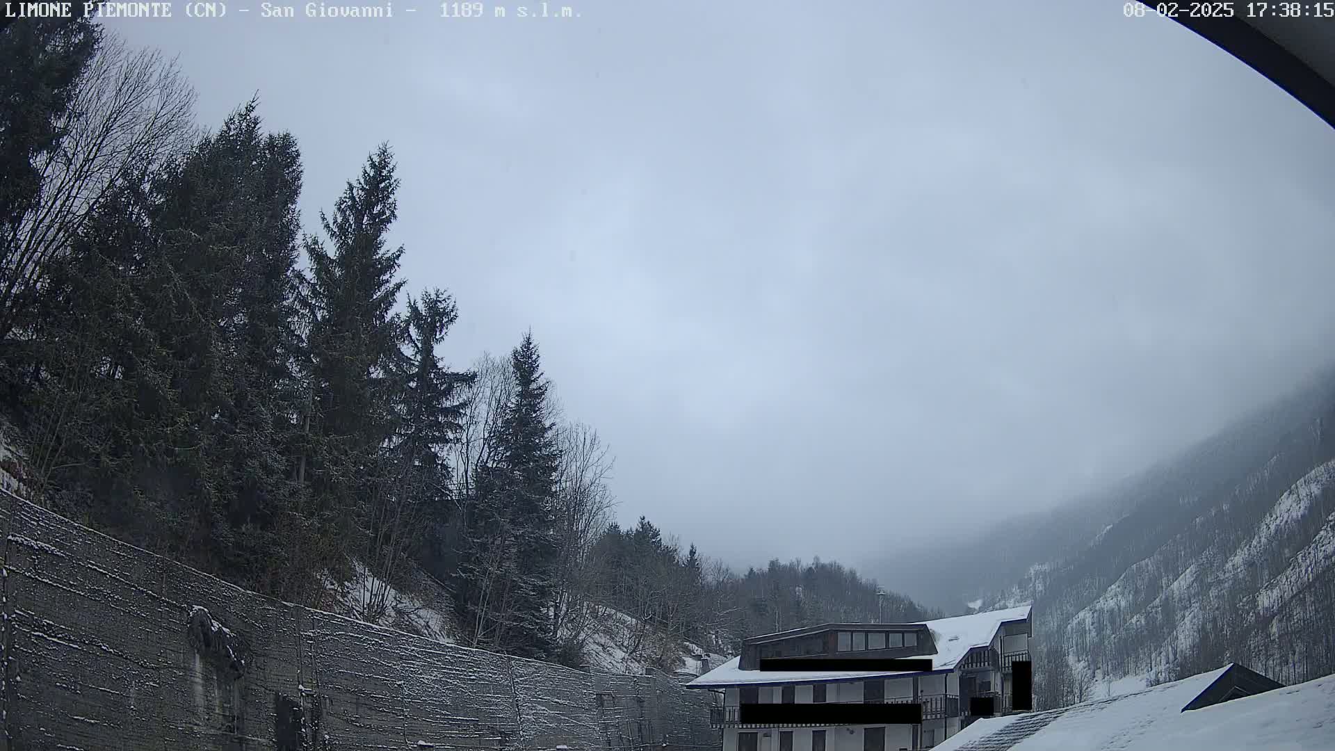 A snow-covered mountain valley scene under an overcast sky shows evergreen trees and a building partially obscured by fog.