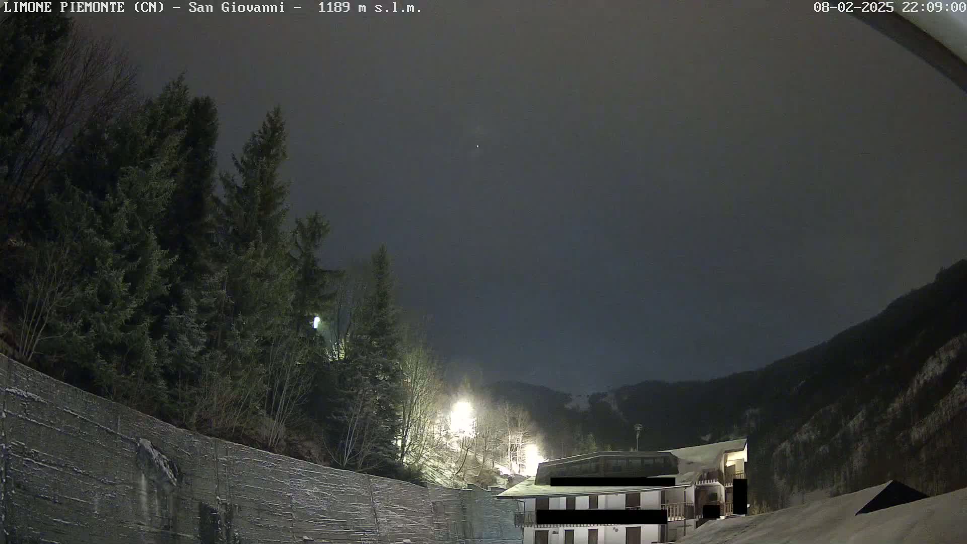 A nighttime view of a snow-covered mountain slope with evergreens, a building, and a retaining wall under a dark, overcast sky.