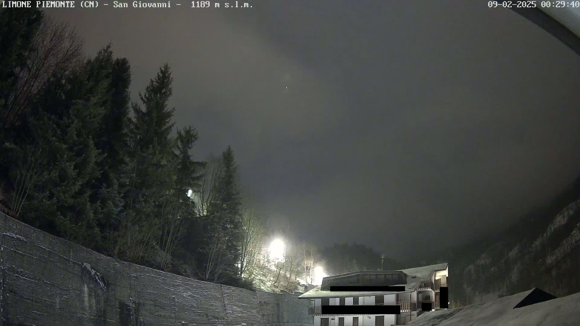A nighttime view of a snow-covered hillside with evergreen trees and a building under a dark, cloudy sky.