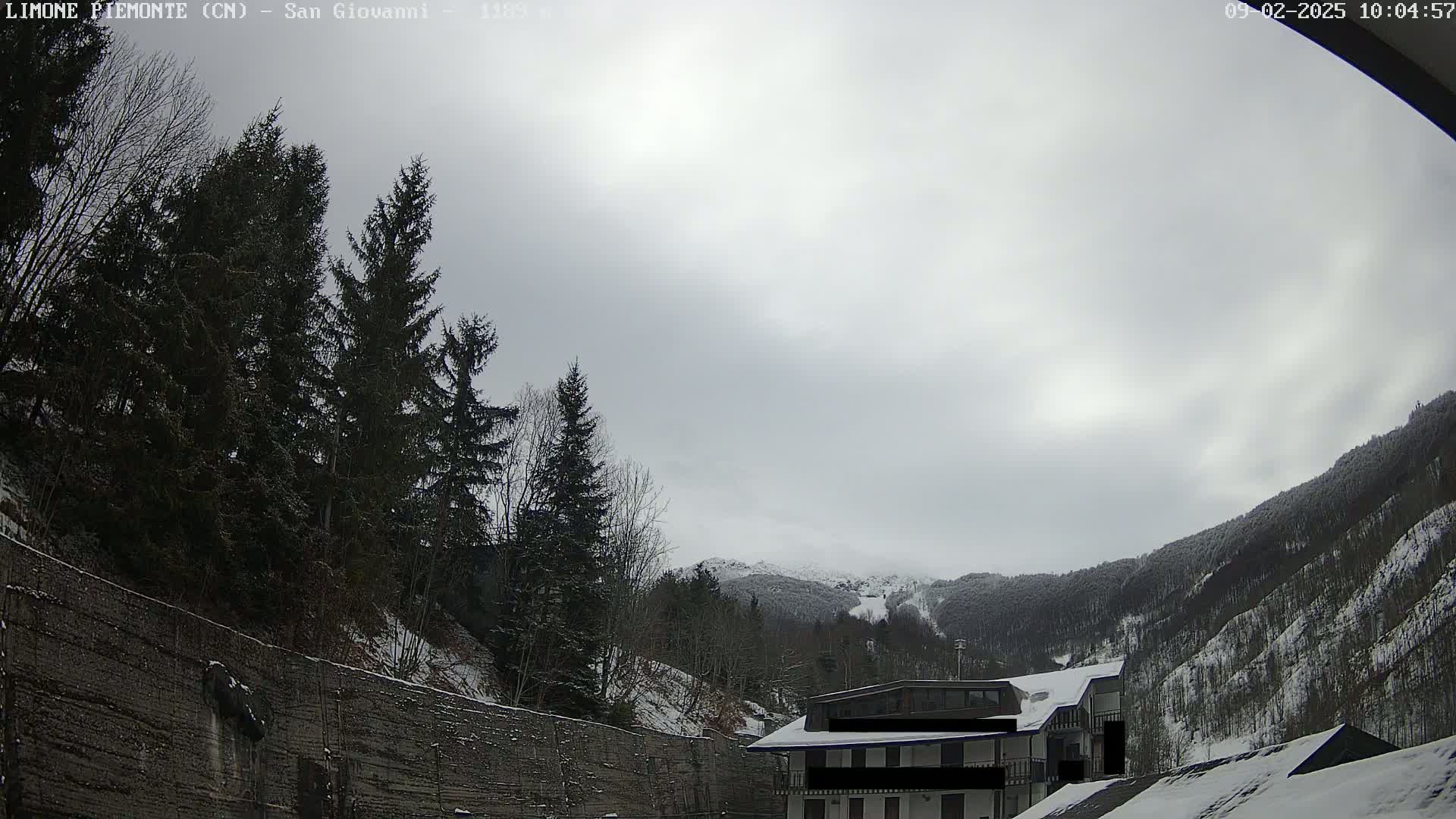 A snow-covered mountainside is visible beyond a building and a wall lined with evergreens under an overcast sky.