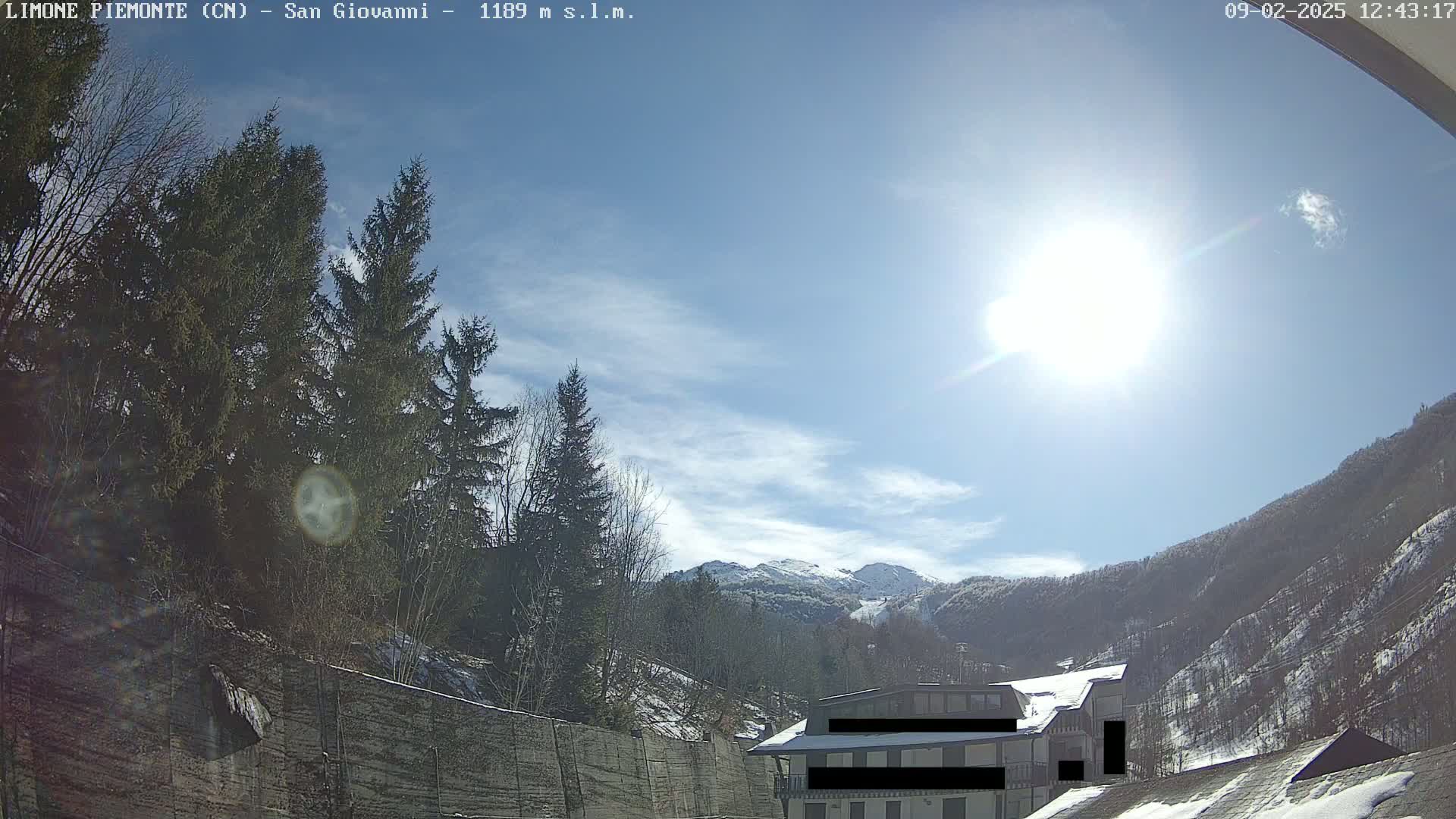 A sunny day in a mountainous region shows a building partially obscured by evergreens and a snow-covered mountain in the background.