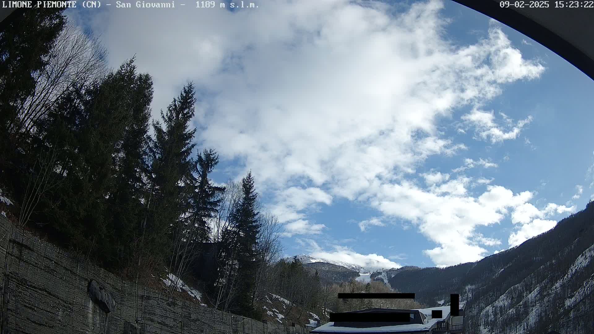 A partly cloudy blue sky is visible above a mountainside covered in snow and evergreen trees, partially obscured by a rocky embankment.