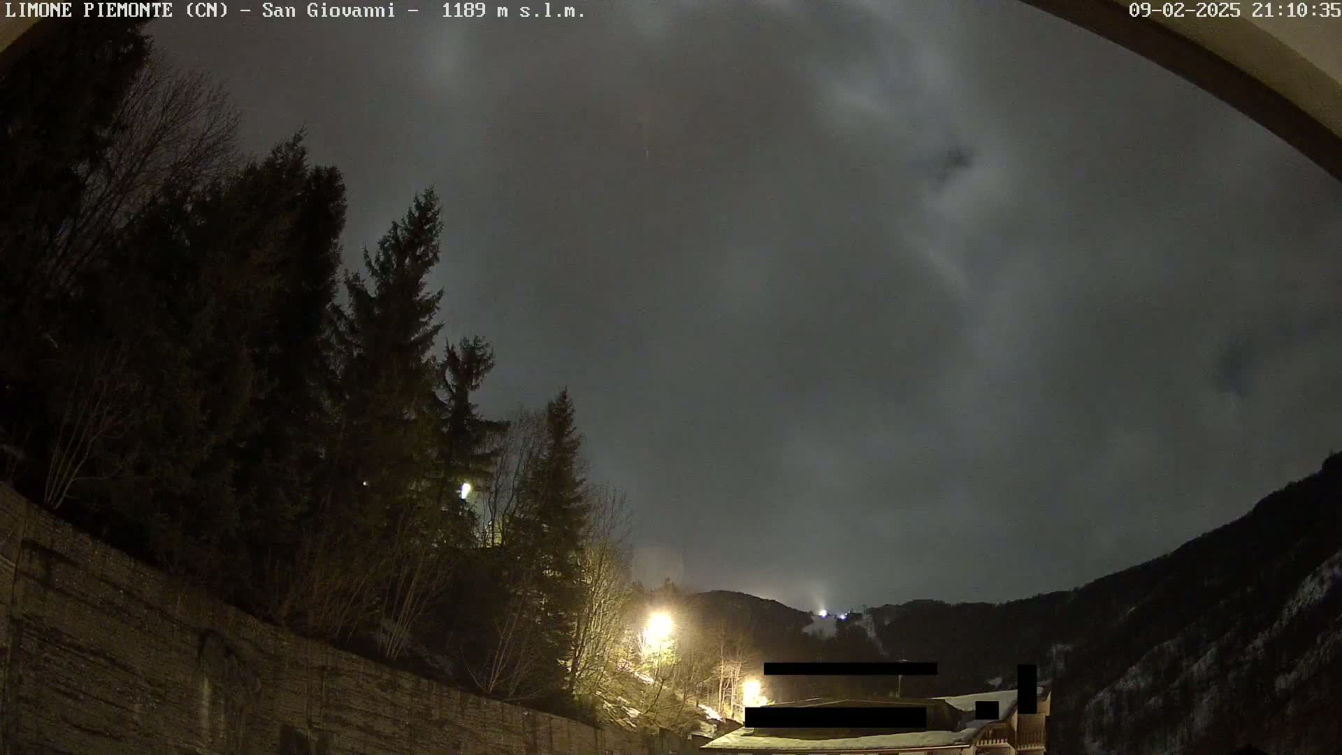 A nighttime view of a snow-covered mountainside under a cloudy sky, with dark evergreen trees and illuminated buildings in the foreground.