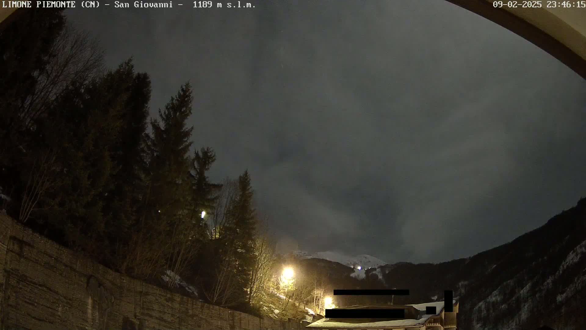 A nighttime view shows a snow-covered mountain in the distance under a cloudy sky, with evergreen trees and a stone wall in the foreground illuminated by streetlights.