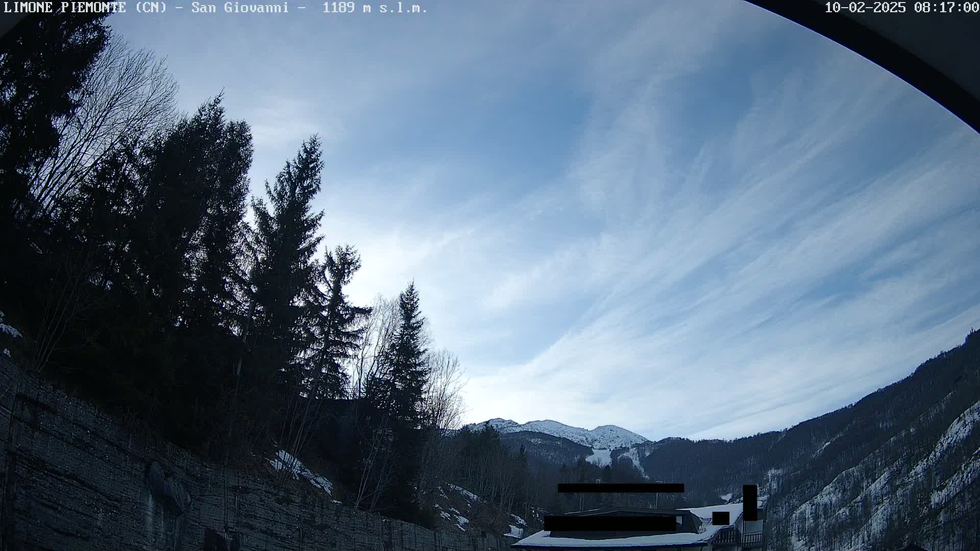 A mostly clear, blue sky with thin, wispy clouds stretches over a snow-dusted mountain range and dark evergreen trees lining a rocky hillside.