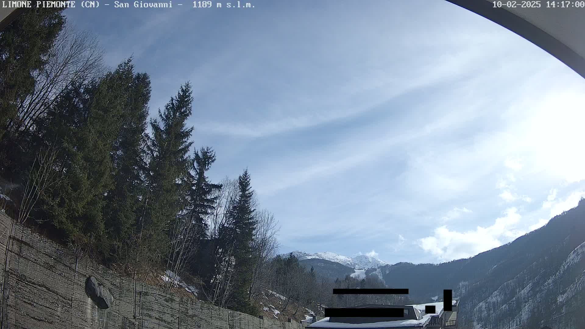 A partly cloudy day reveals snow-capped mountains in the distance, viewed from a roadway lined with evergreens and a retaining wall.