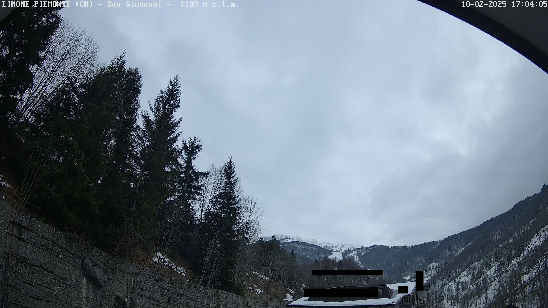 A cloudy sky hangs over snow-dusted mountains and evergreens, seen from within a vehicle near a tall, stone retaining wall.