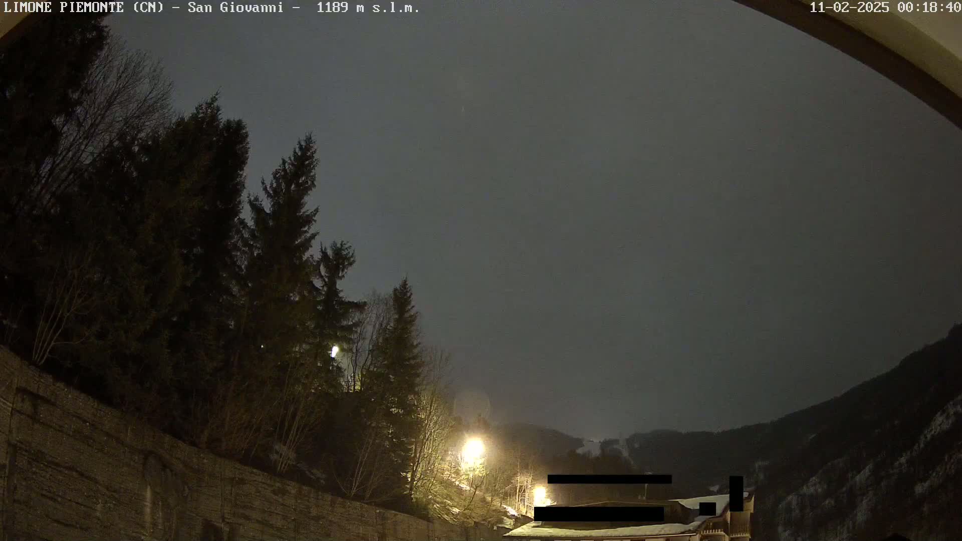 A nighttime view of a snow-covered mountainside under an overcast sky, illuminated by streetlights, with dark evergreen trees in the foreground.