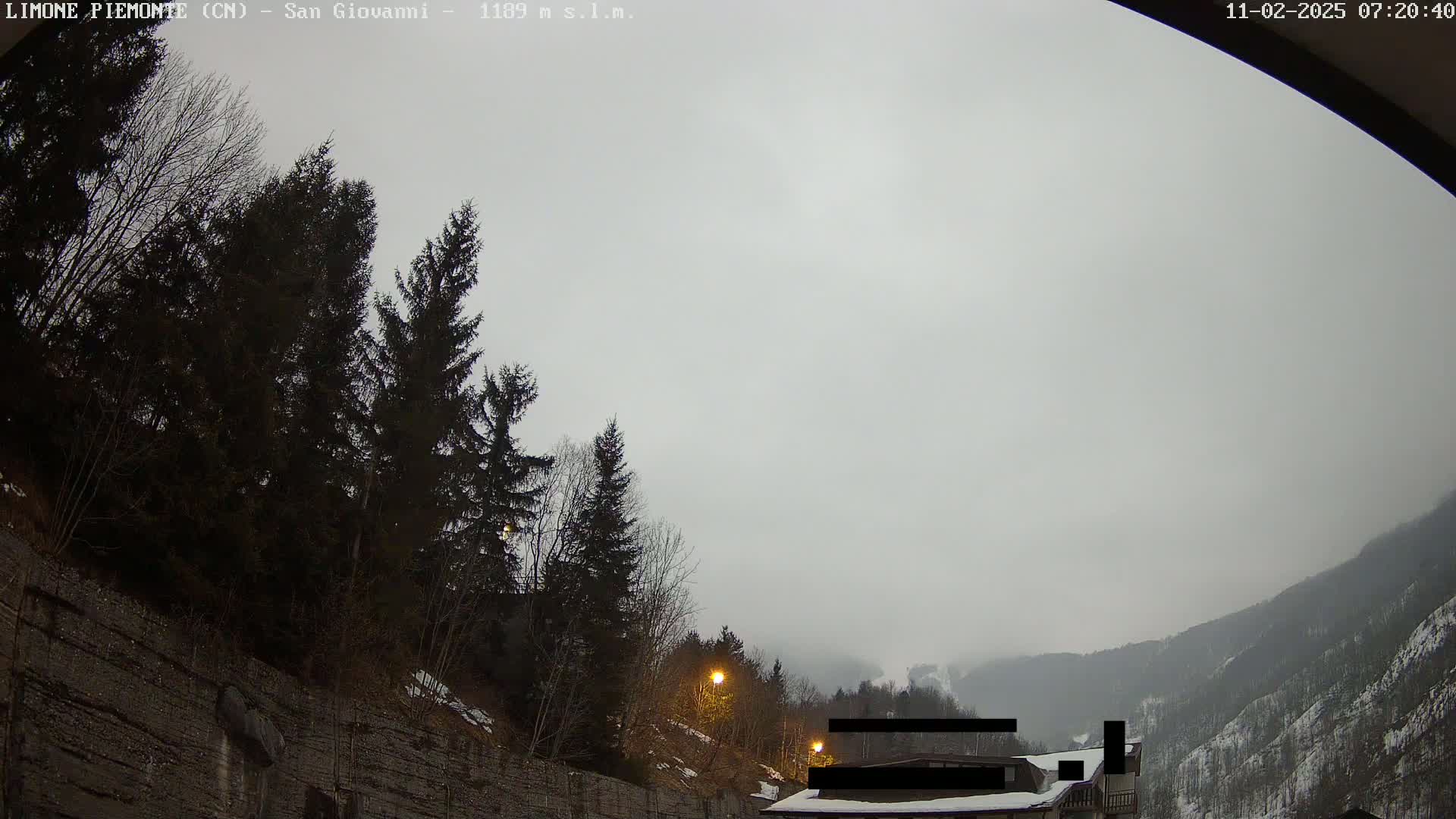 A view of a mountainous area under an overcast sky shows dark evergreens next to a rocky retaining wall, with a building partially visible in the distance.