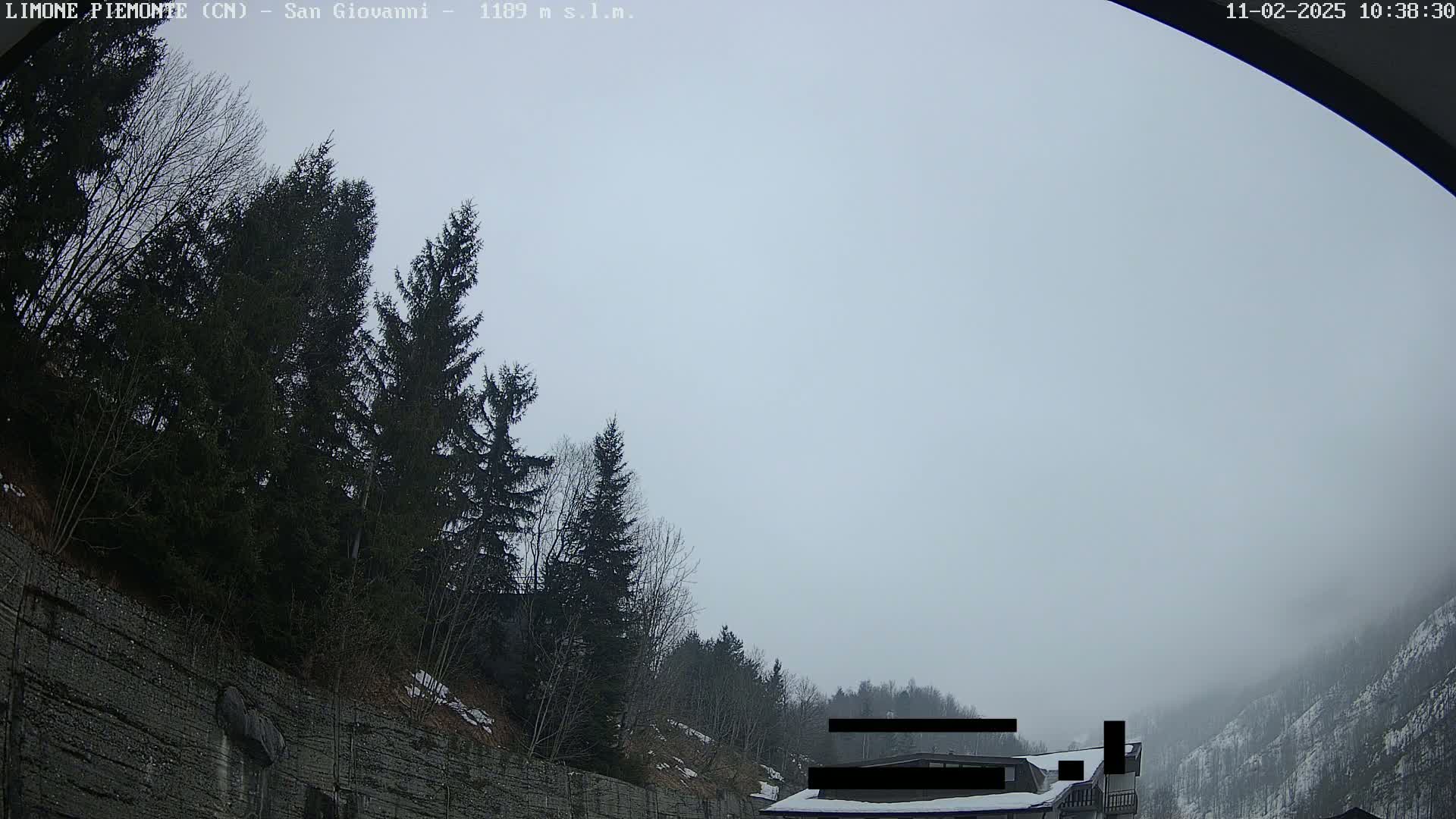 A foggy, overcast day reveals a mountainous landscape with evergreen trees and a stone retaining wall, partially obscuring a building in the distance.