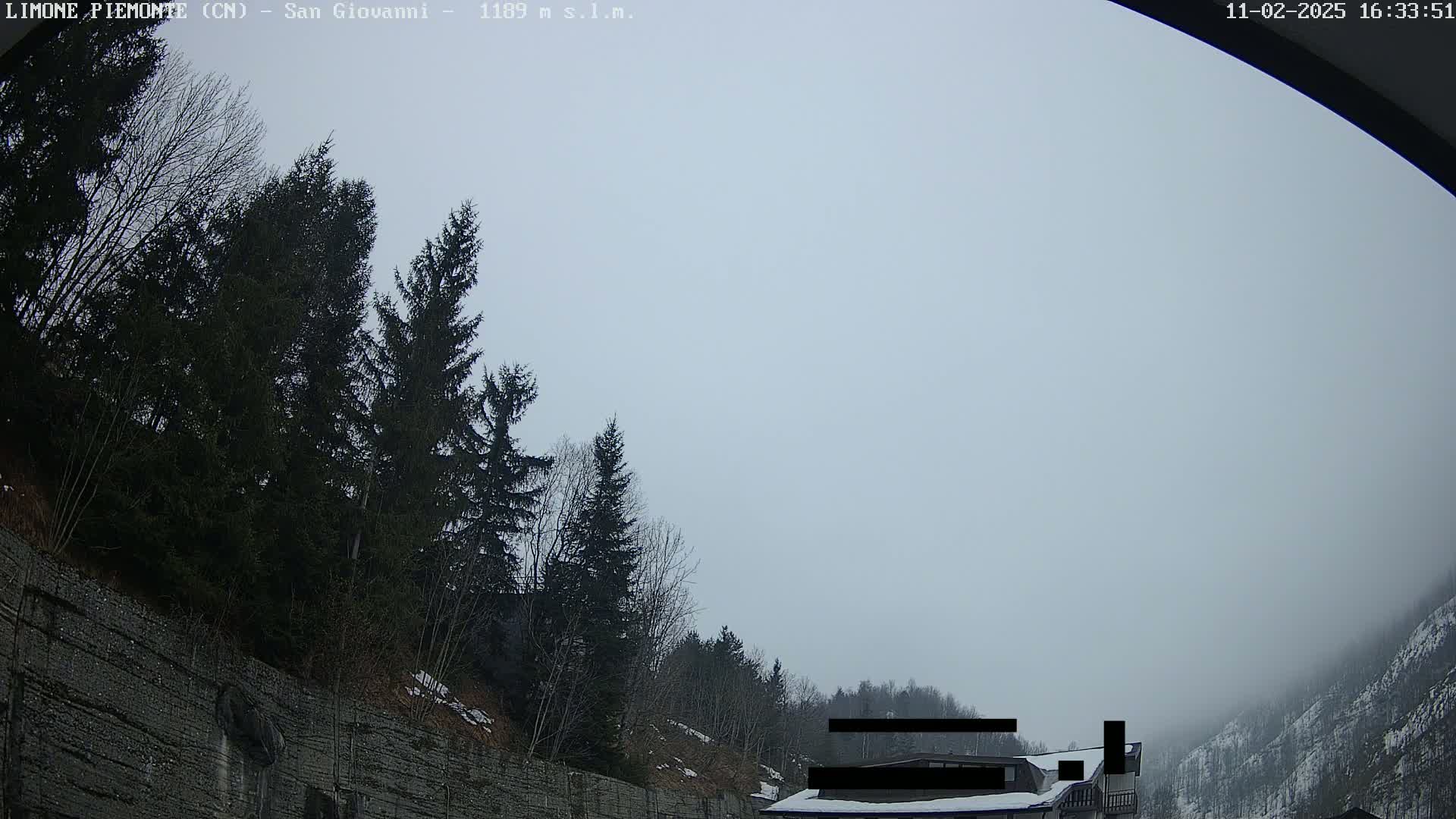 A mostly overcast sky hangs over a mountainous landscape with dark evergreens growing along a stone wall, and a building partially obscured by fog in the distance.