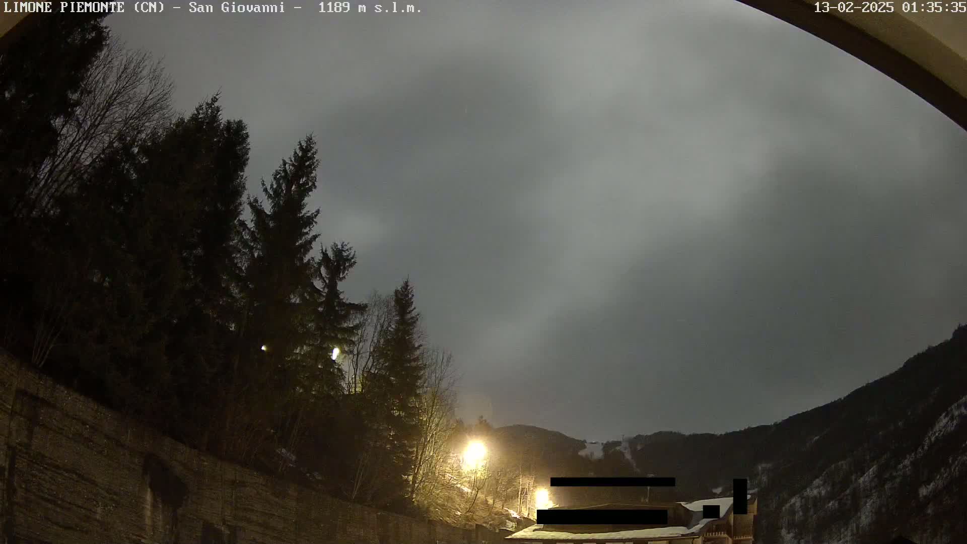 Under an overcast night sky, illuminated streetlights highlight evergreen trees and a mountainside partially covered in snow.