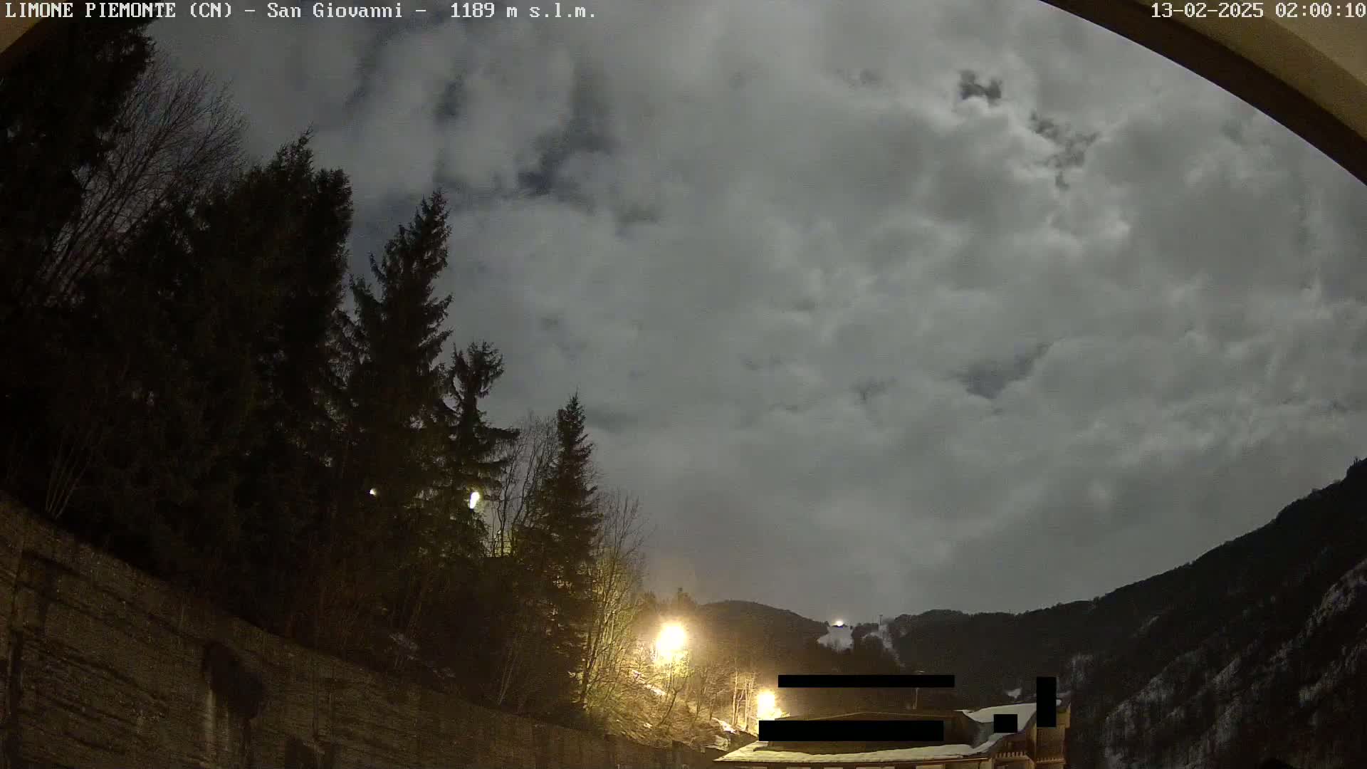 A nighttime view of a mountainside with a cloudy sky, illuminated by streetlights, showing dark evergreen trees and a stone wall in the foreground.
