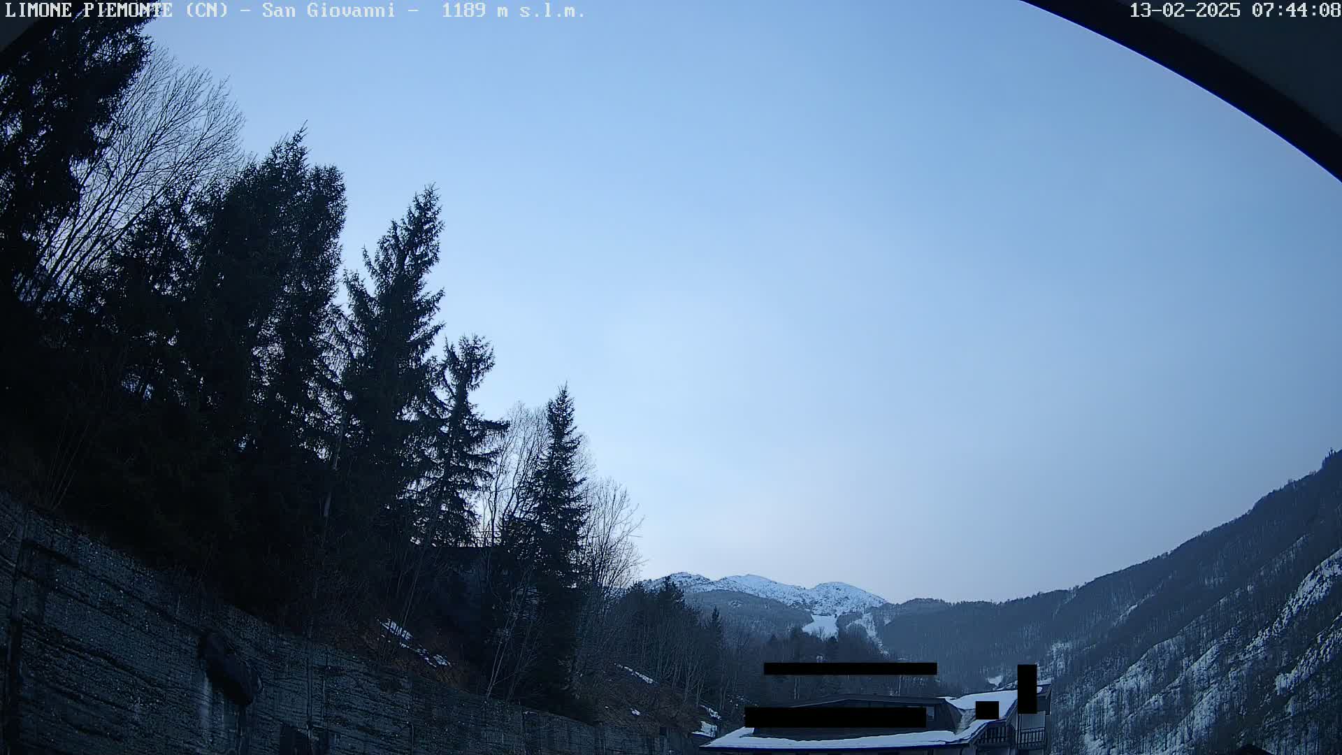 A clear, pale blue sky overlooks a mountainous landscape with snow-capped peaks in the distance, partially obscured by dark evergreen trees and a stone wall in the foreground.