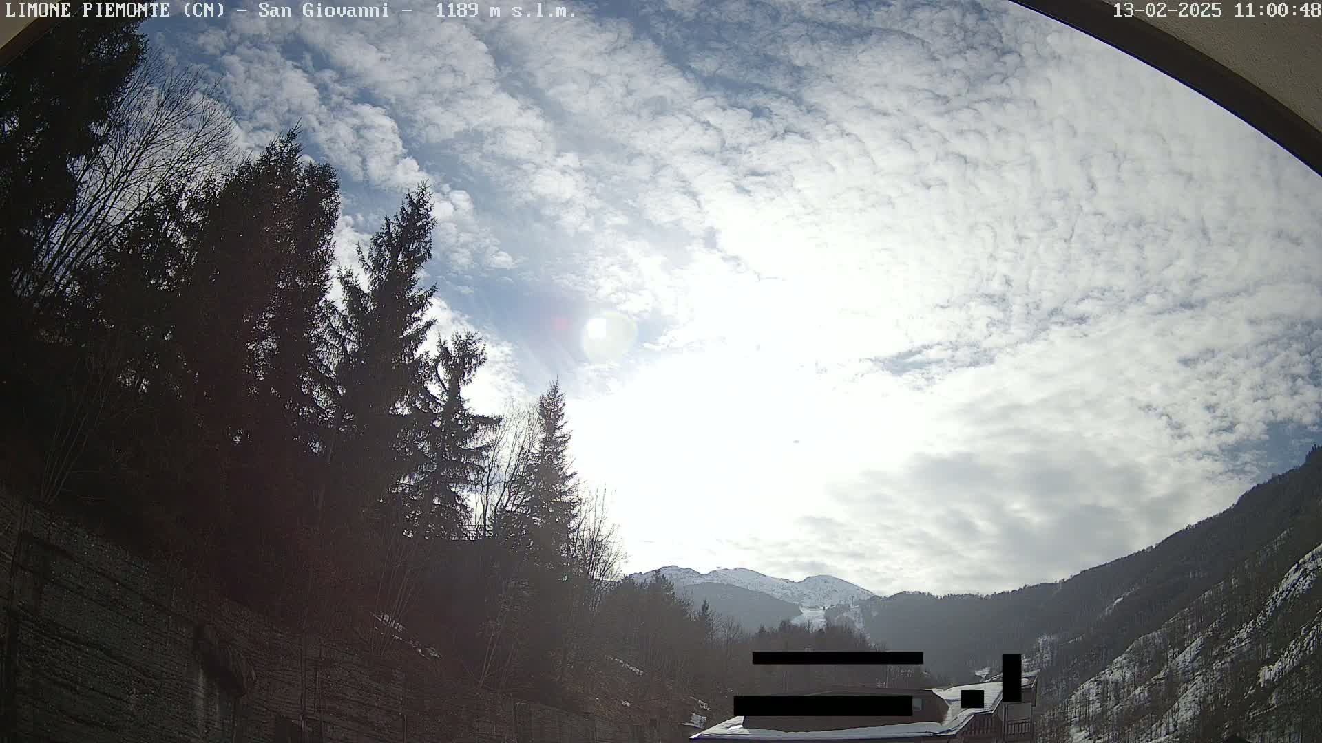 A mostly sunny day with scattered clouds is seen from a viewpoint overlooking snow-capped mountains and a forest of dark evergreen trees.