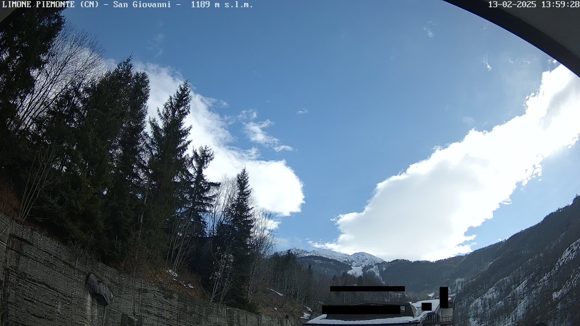 A partly cloudy sky is visible above a mountainous landscape with evergreen trees and a stone wall, some of which is covered in snow.