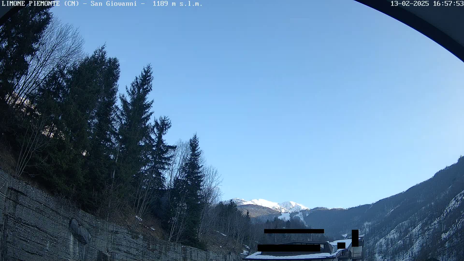 A clear blue sky is visible above a mountain valley with evergreen trees and snow-capped mountains in the distance.
