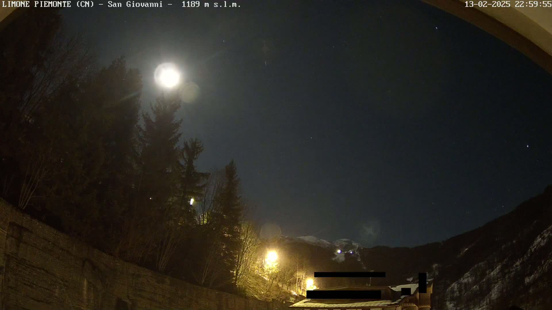 A bright moon illuminates a nighttime scene of evergreen trees, a stone wall, and distant snow-capped mountains under a clear, starry sky.