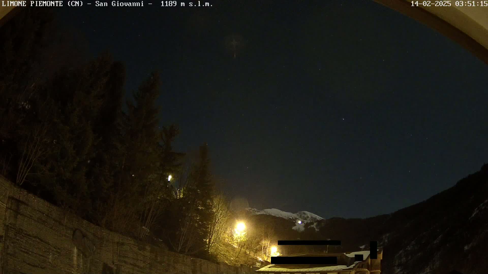 A nighttime view of a snow-capped mountain in the distance, behind a dark, tree-lined hillside illuminated by streetlights.