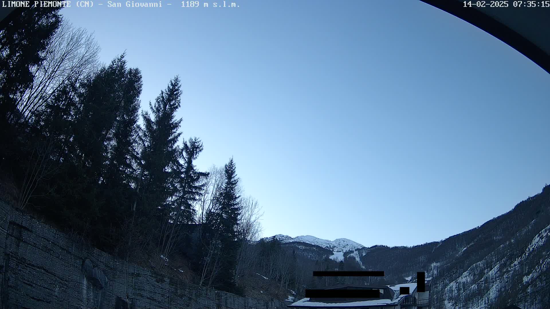 A clear blue sky is visible above a snow-capped mountain and dark evergreen trees, some of which are situated on a steep, rocky hillside.