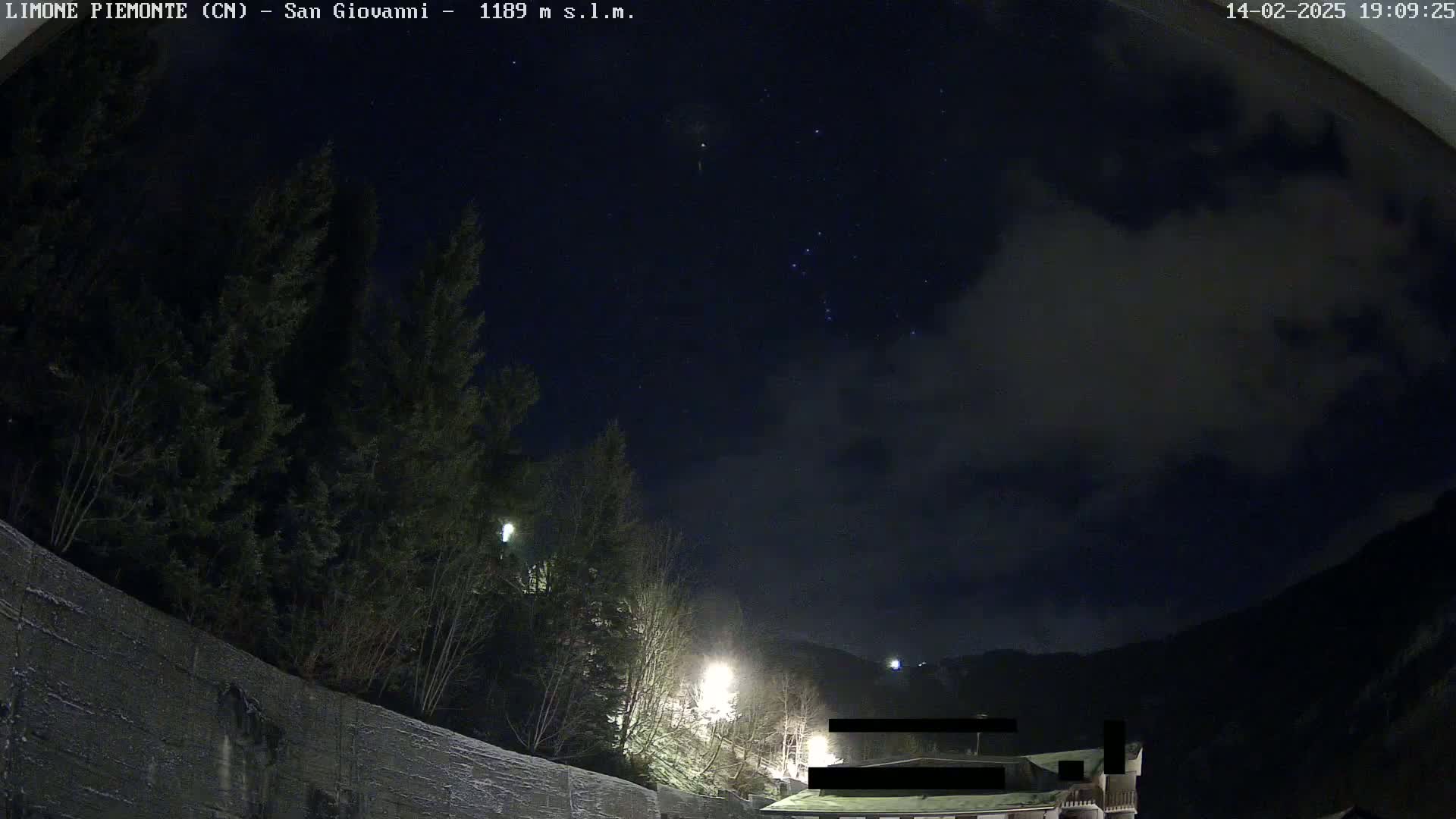 A nighttime view of a snow-covered landscape, featuring evergreens, streetlights, and a partly cloudy sky with some stars visible.