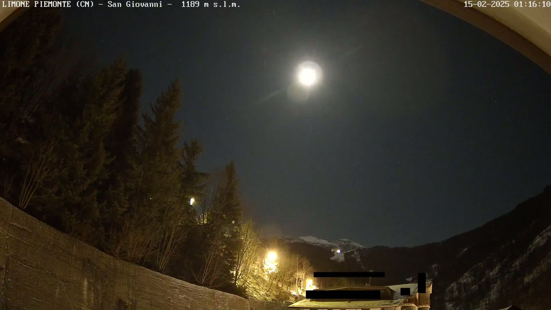 A bright moon illuminates a nighttime scene of snow-covered mountains and evergreens, with some lights visible near buildings in the foreground.