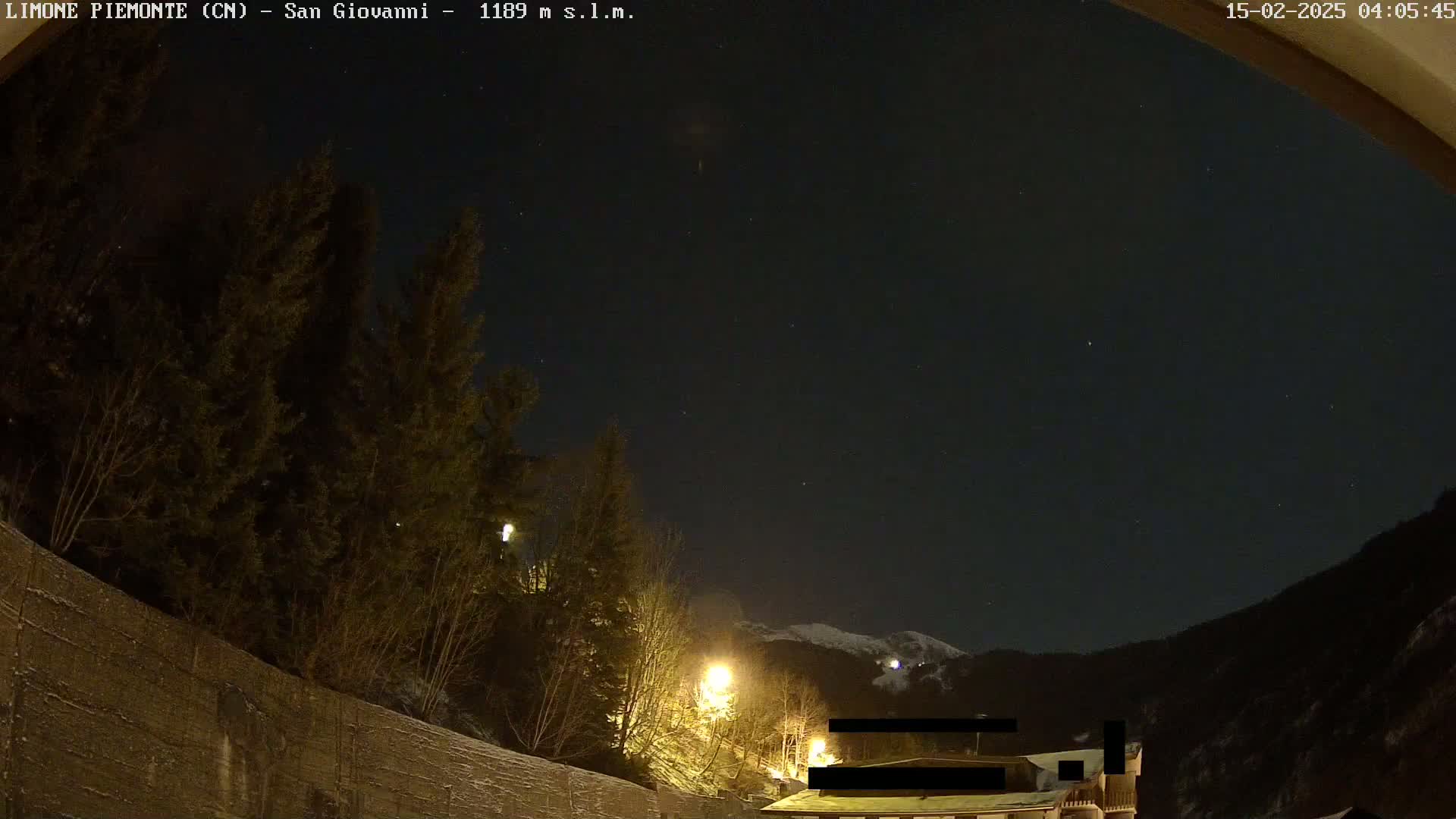 A nighttime view of a snow-covered mountainside, illuminated by streetlights, with a clear, starry sky above and dark evergreen trees in the foreground.