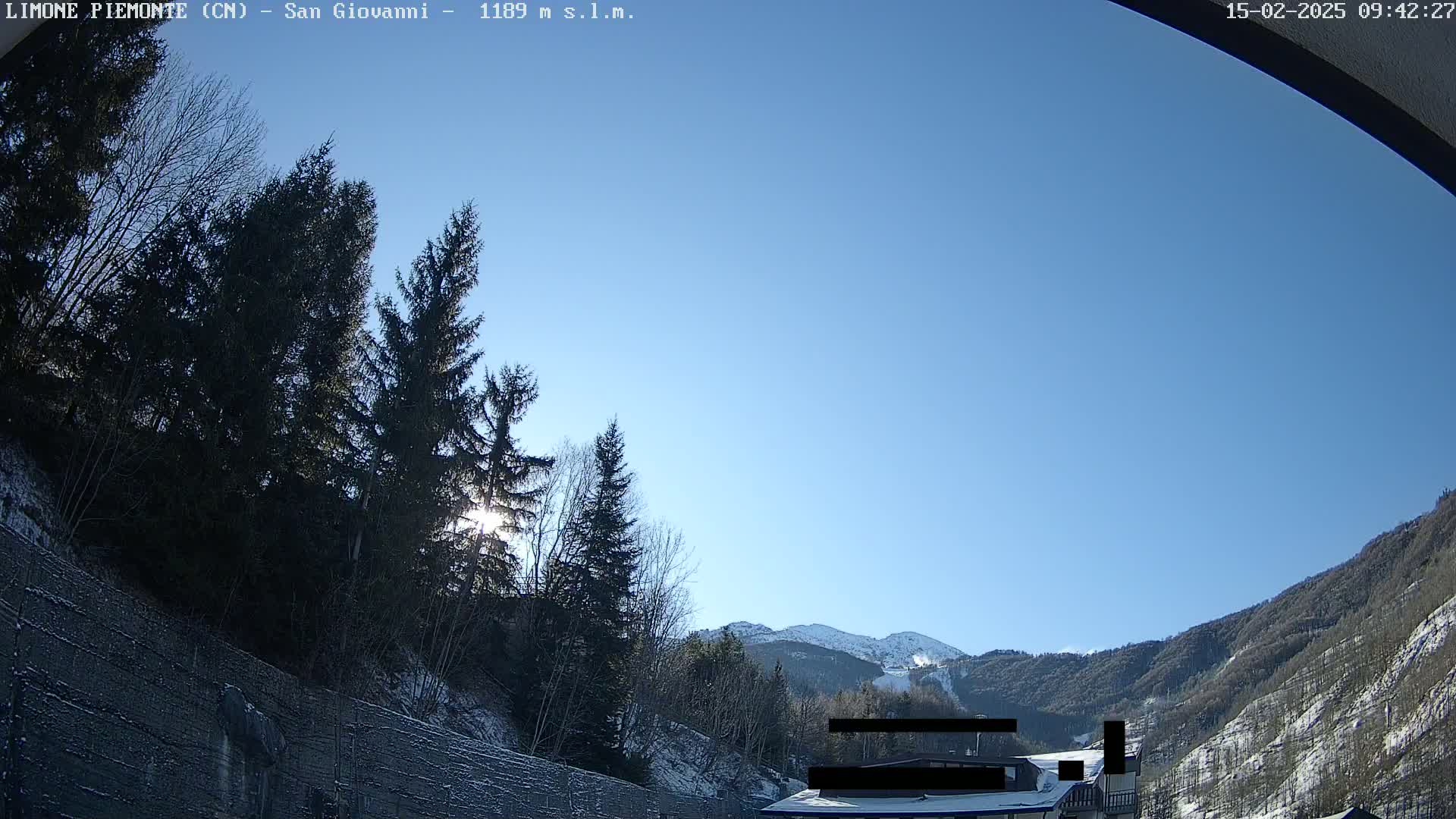 A sunny, clear day in a snow-dusted mountain valley, viewed from behind a tall concrete wall, shows evergreen trees and a partially visible building in the foreground.