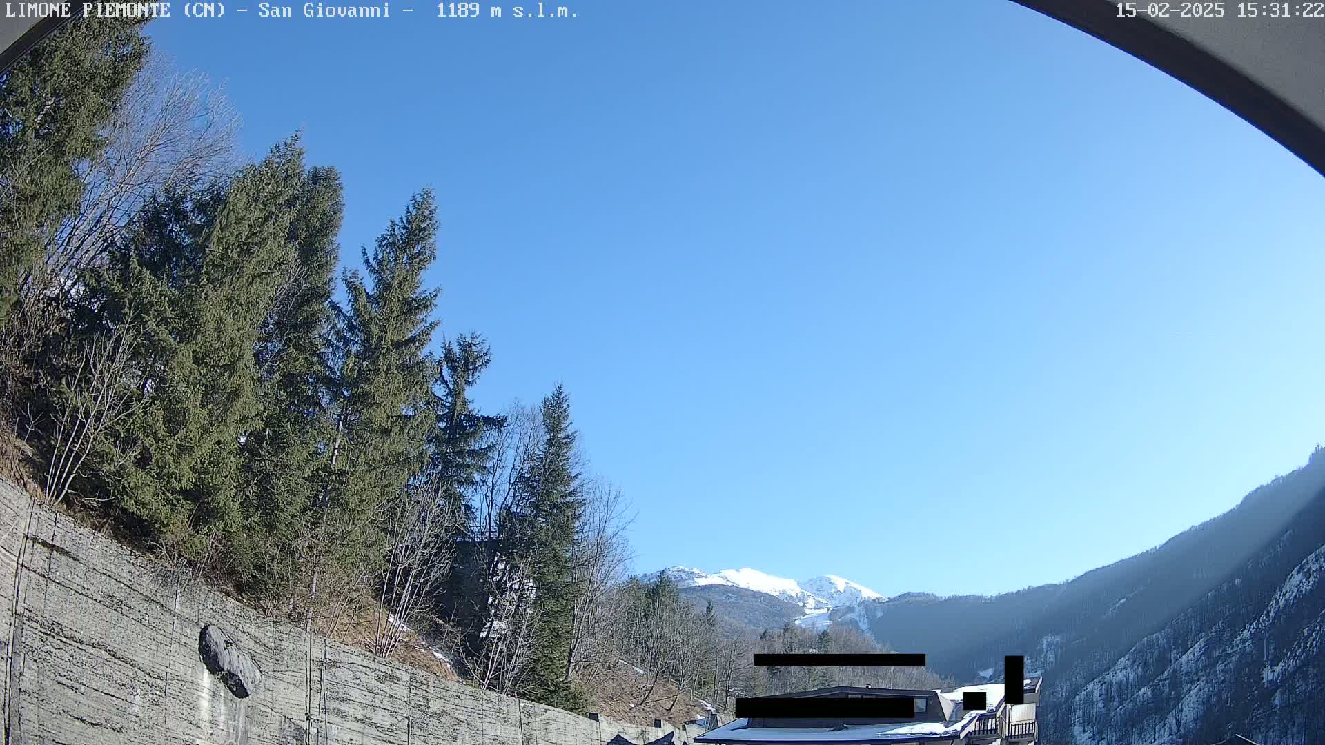 A clear blue sky overlooks a mountainous landscape with snow-capped peaks in the distance, partially obscured by evergreens and a concrete retaining wall.