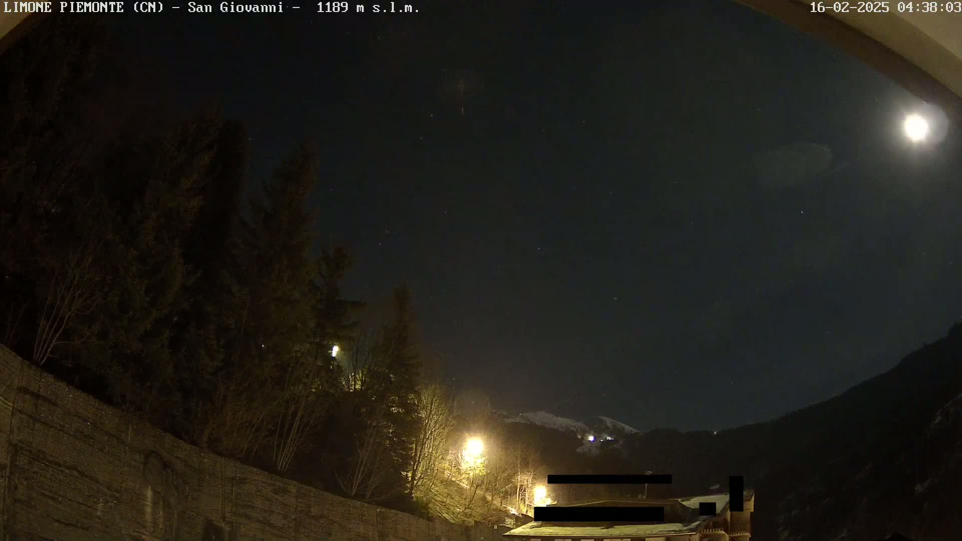 A nighttime view shows a moonlit sky above dark trees and a stone wall, with a few lights illuminating the scene and snow-covered mountains in the distance.