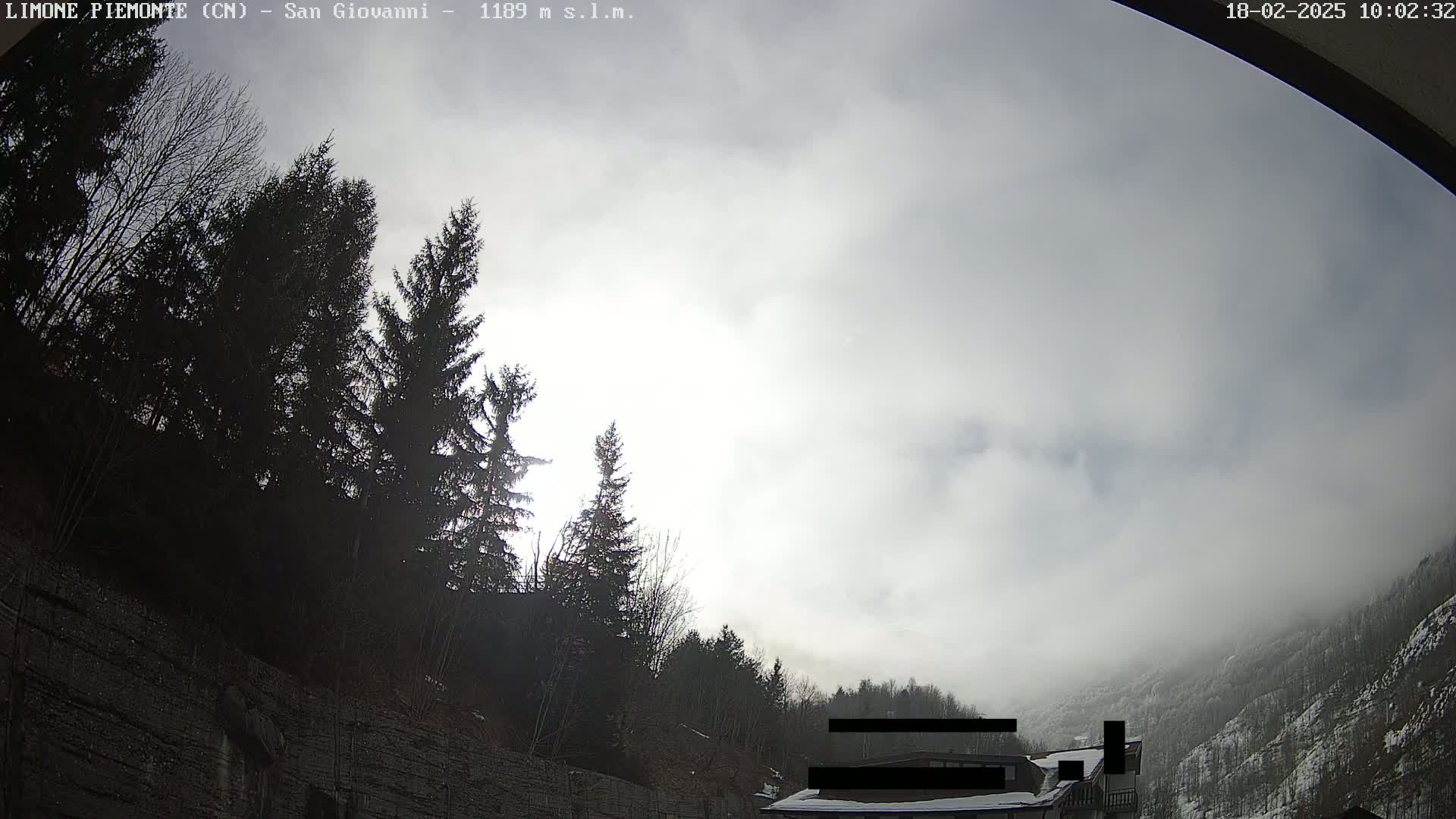 A mostly cloudy sky with some sunlight breaking through, overlooking a hillside of dark evergreen trees and a building partially obscured by fog.
