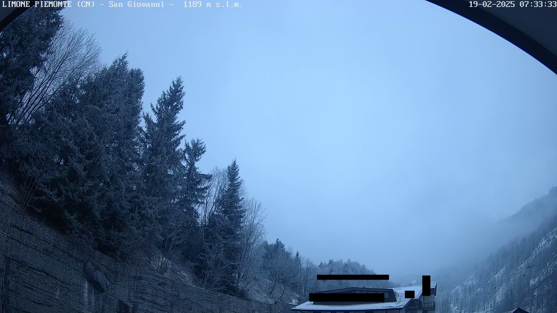 A hazy, overcast sky hangs over a snow-dusted mountainside with evergreen trees and a partially visible building.