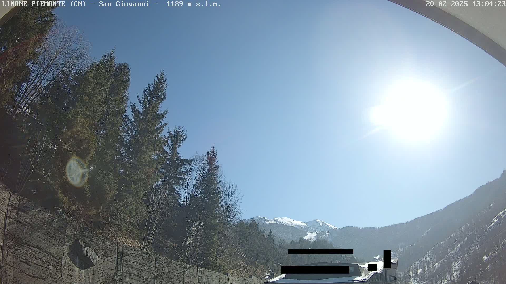 A sunny day in a mountainous area shows evergreens and a snow-capped mountain in the distance, viewed from behind a low concrete wall.
