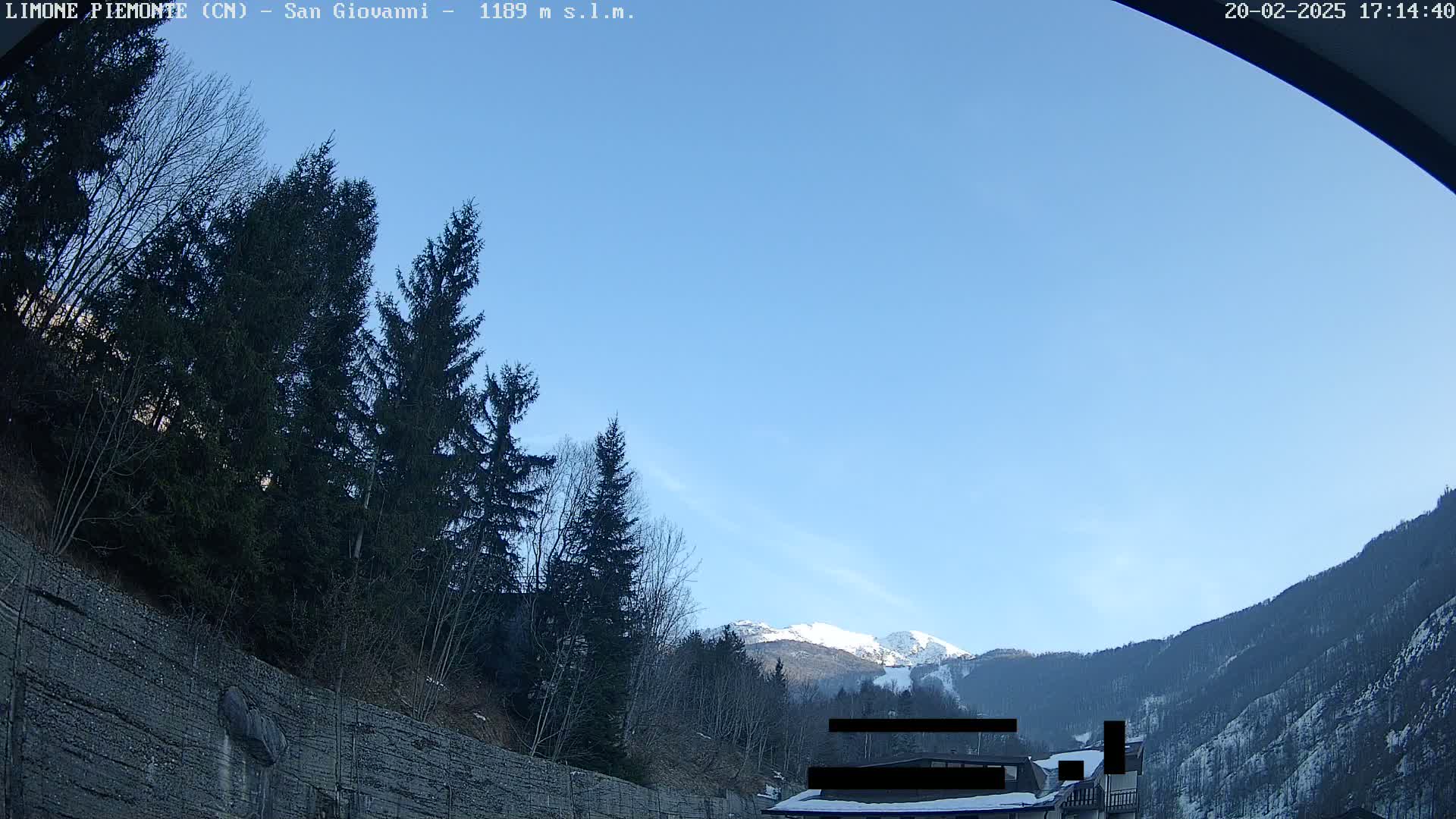 A clear blue sky overlooks a mountain range partially covered in snow, with evergreens in the foreground and a rocky retaining wall.