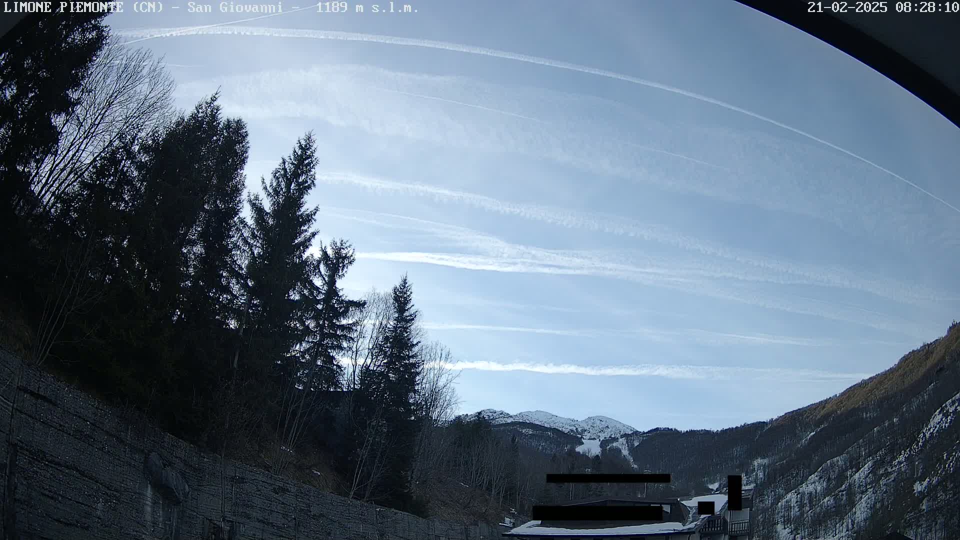 A mostly clear, bright blue sky with contrails shows above a mountain range partially covered in snow, with dark evergreen trees in the foreground.