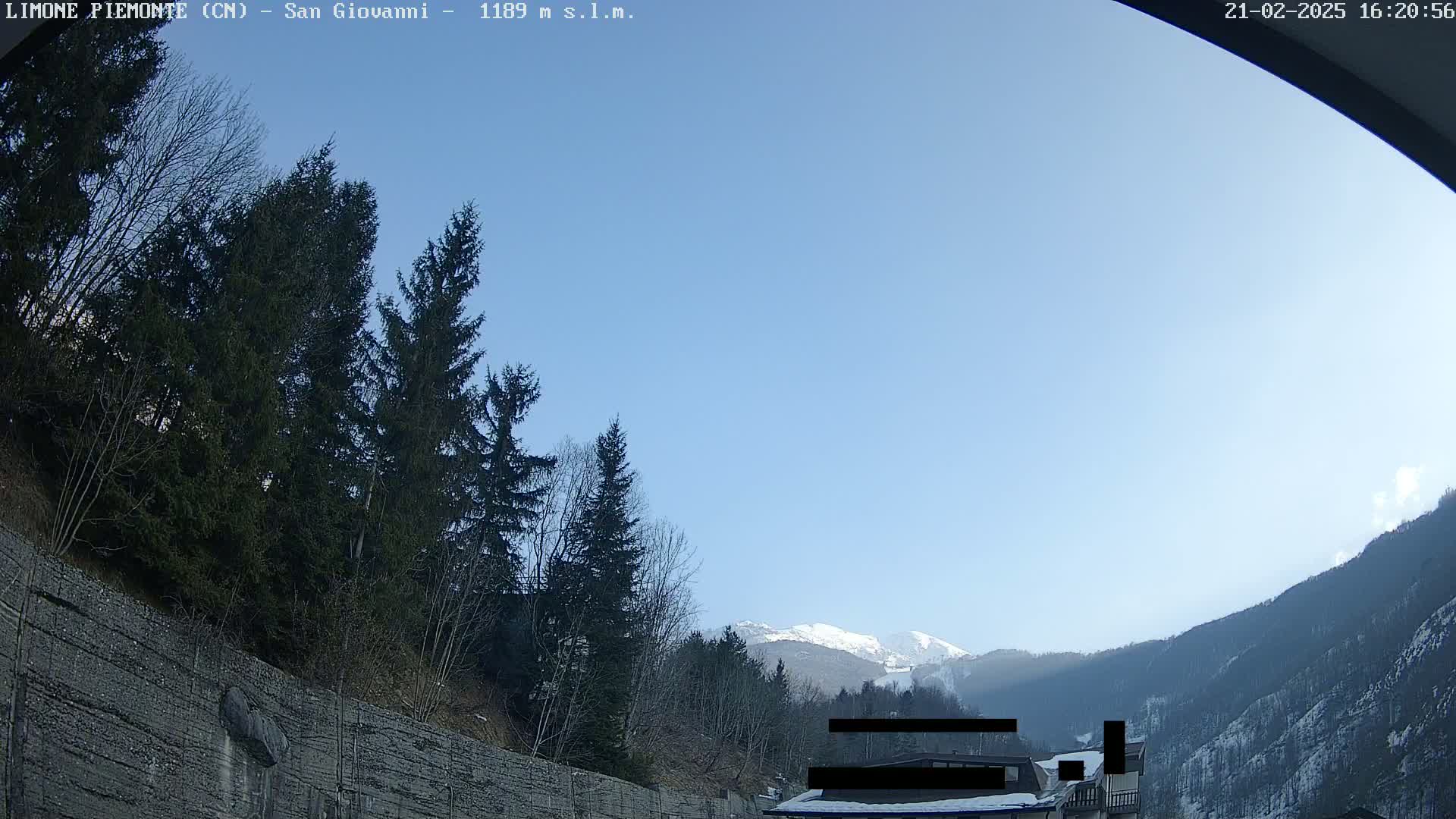 A clear blue sky overlooks snow-capped mountains and evergreen trees, with a rocky retaining wall in the foreground.