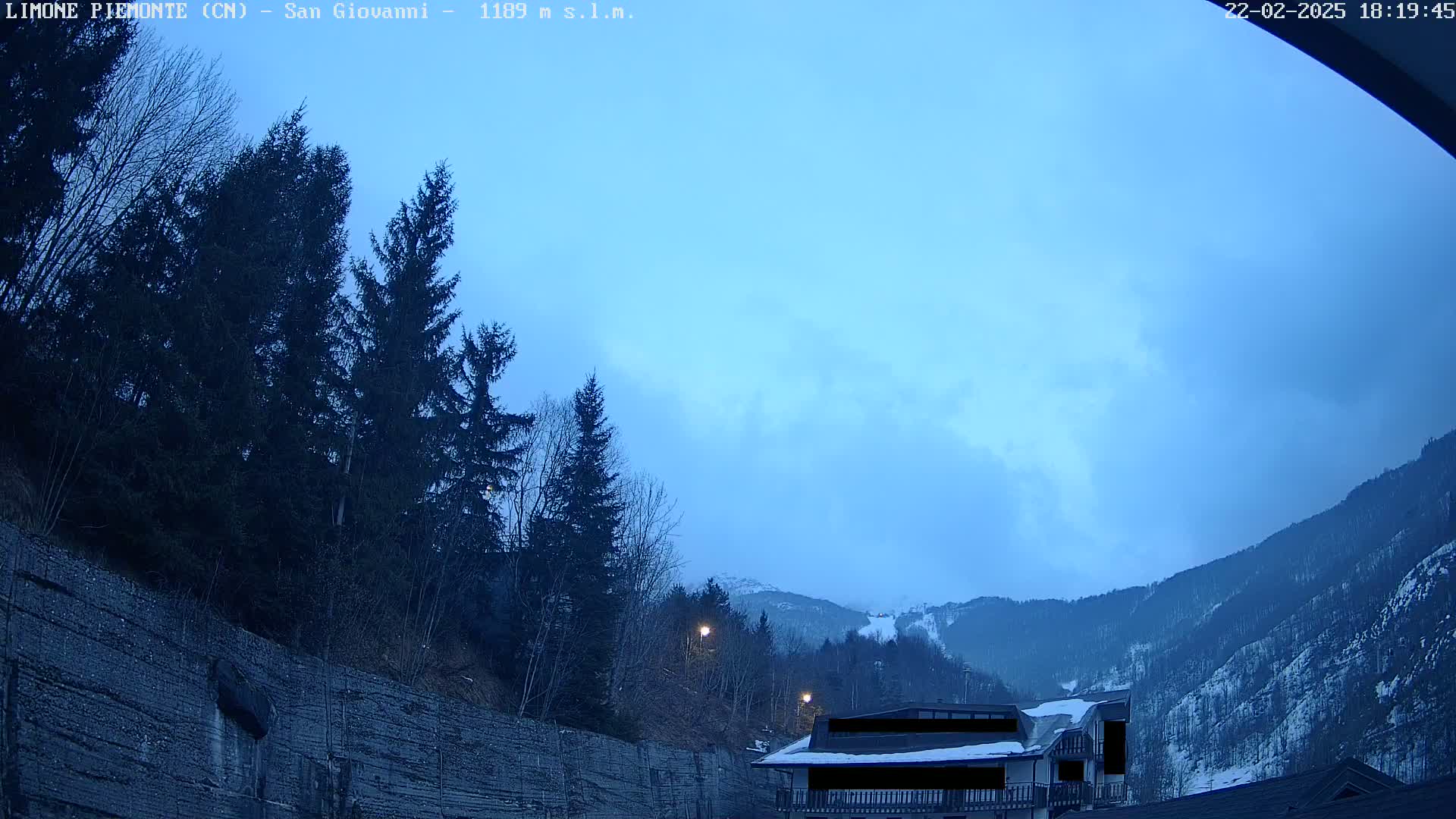 A twilight view shows a snow-dusted mountainside under a mostly clear, dusky blue sky, with evergreens and a building in the foreground.